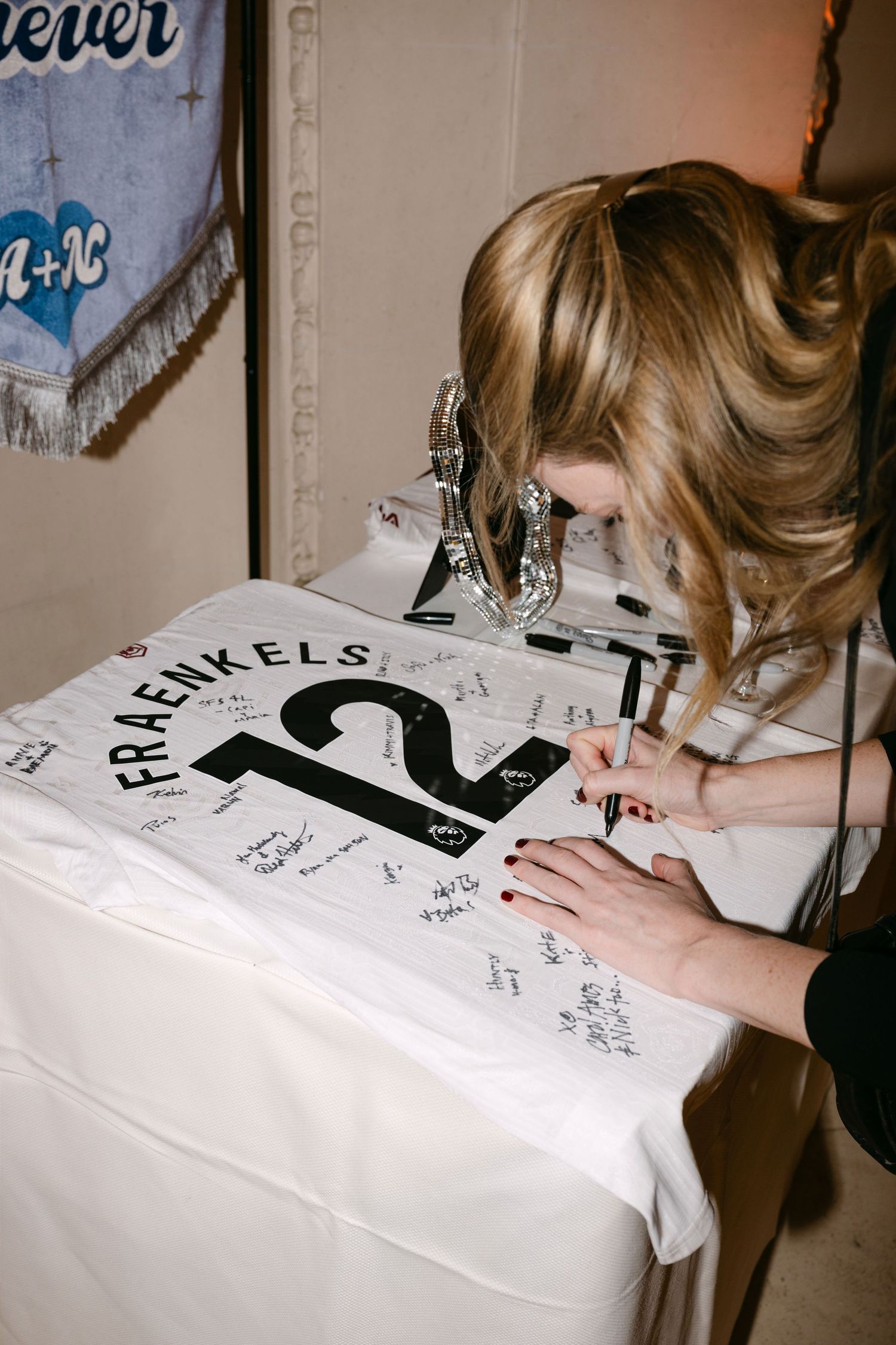 Blonde wedding guest signing sports jersey guest book reading Fraenkels 12 at elegant reception table.