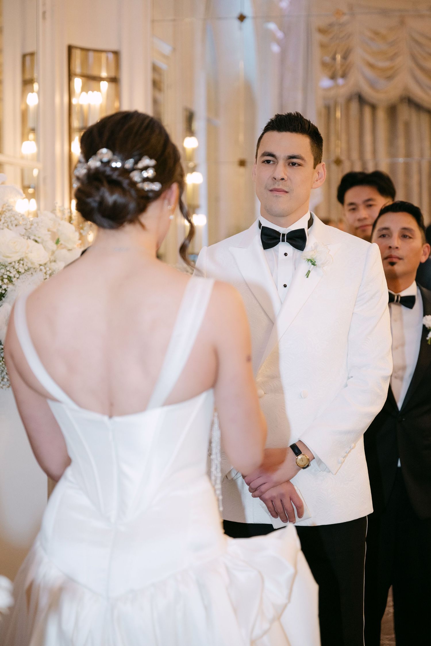 Groom in white tuxedo jacket gazes at bride during elegant indoor wedding ceremony with groomsmen behind him.