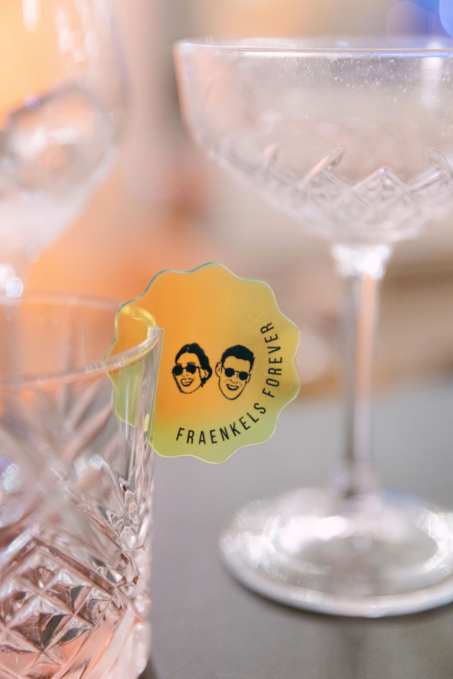 Yellow sticker reading Fraenkels Forever with illustrated faces attached to a crystal glass at a wedding reception.