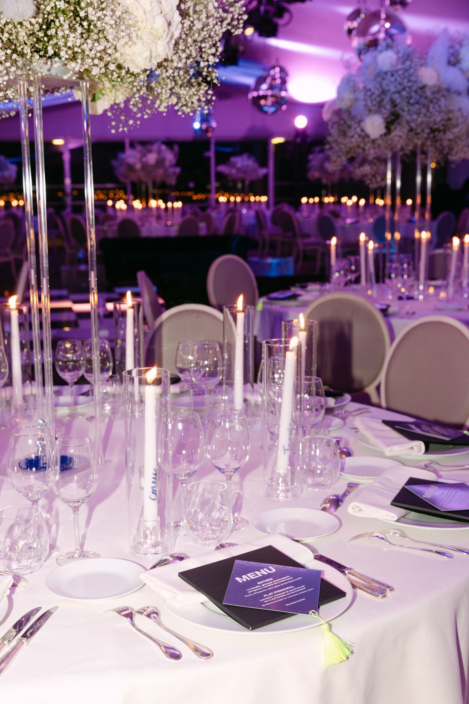 Romantic wedding reception tables with tall floral centerpieces, candles, and purple ambient lighting.