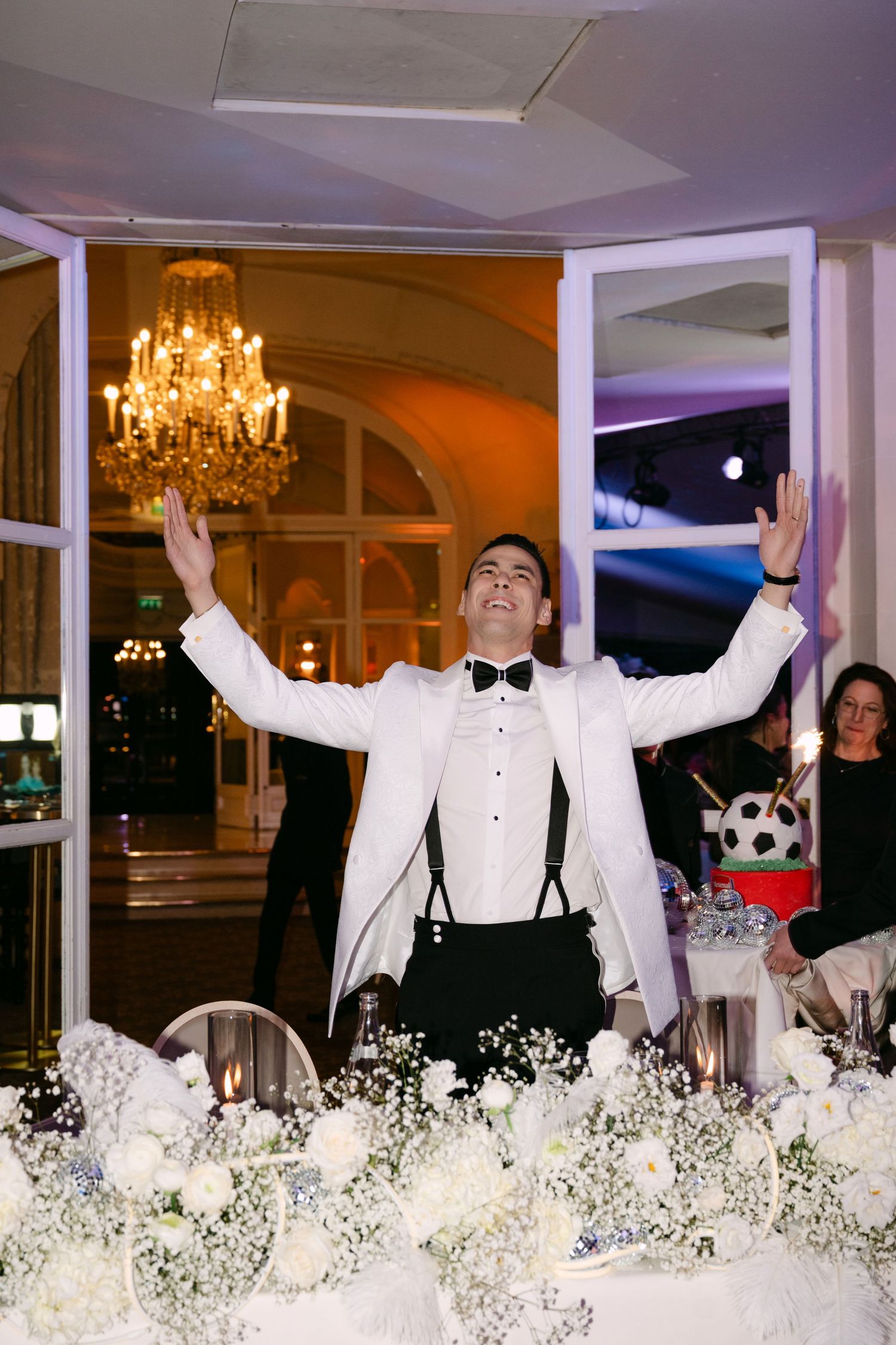 Groom in white tuxedo raises arms triumphantly at elegant wedding reception with chandelier backdrop.