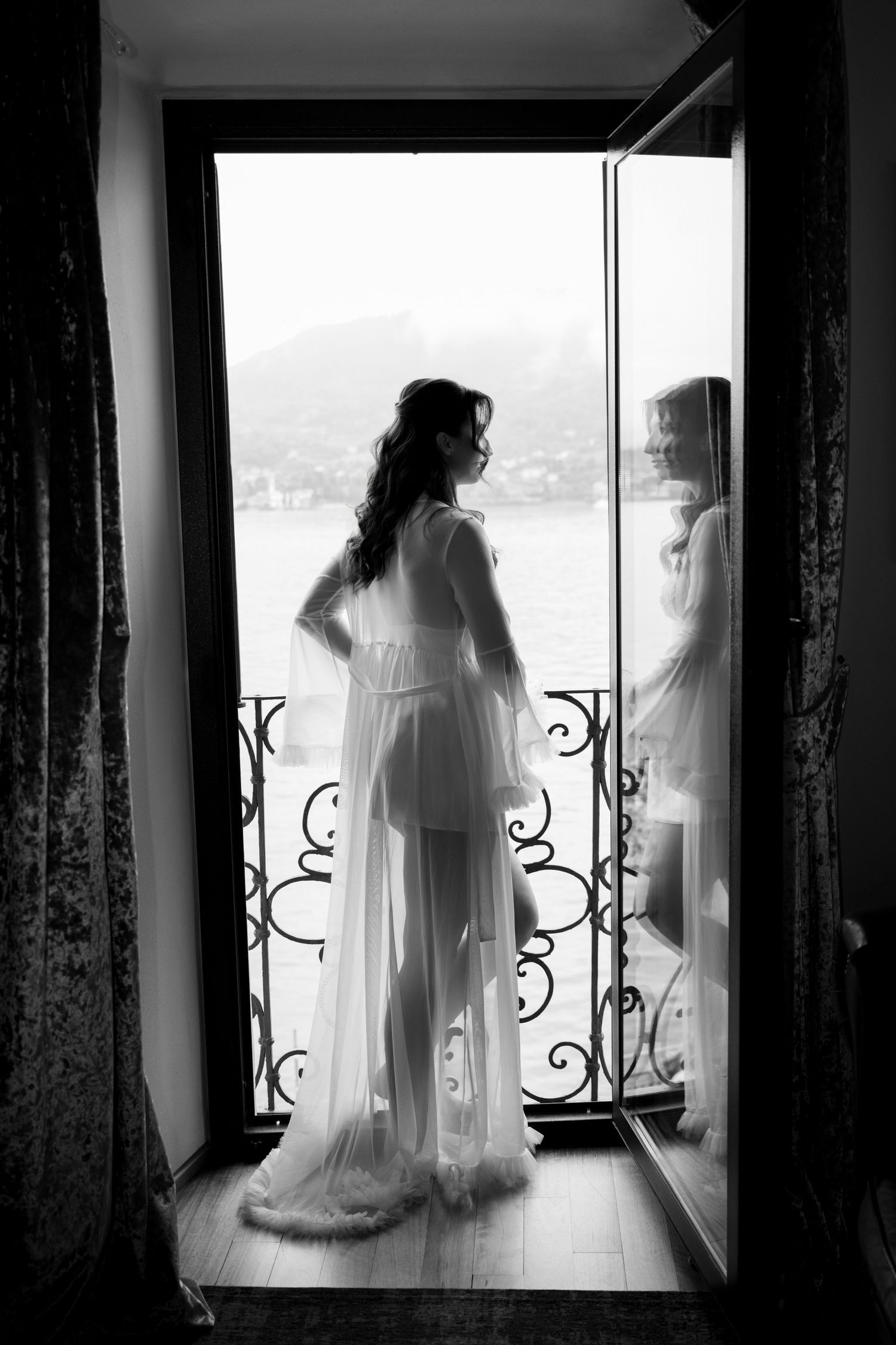 Black and white photo of a bride in a sheer robe standing at an open balcony door overlooking a scenic waterfront.