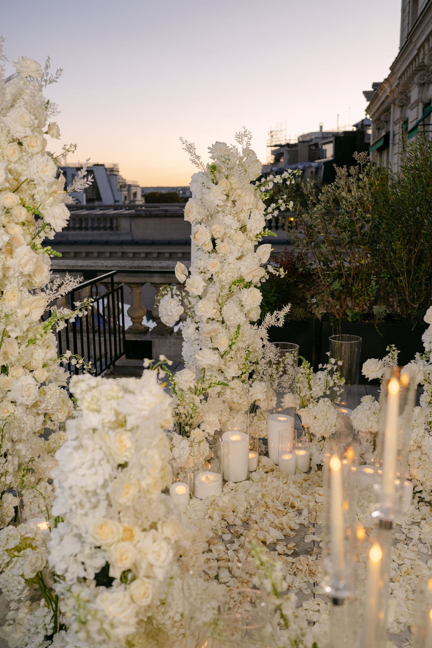 White roses and hydrangeas with glowing candles decorate a Paris rooftop terrace at sunset for a romantic proposal setup.