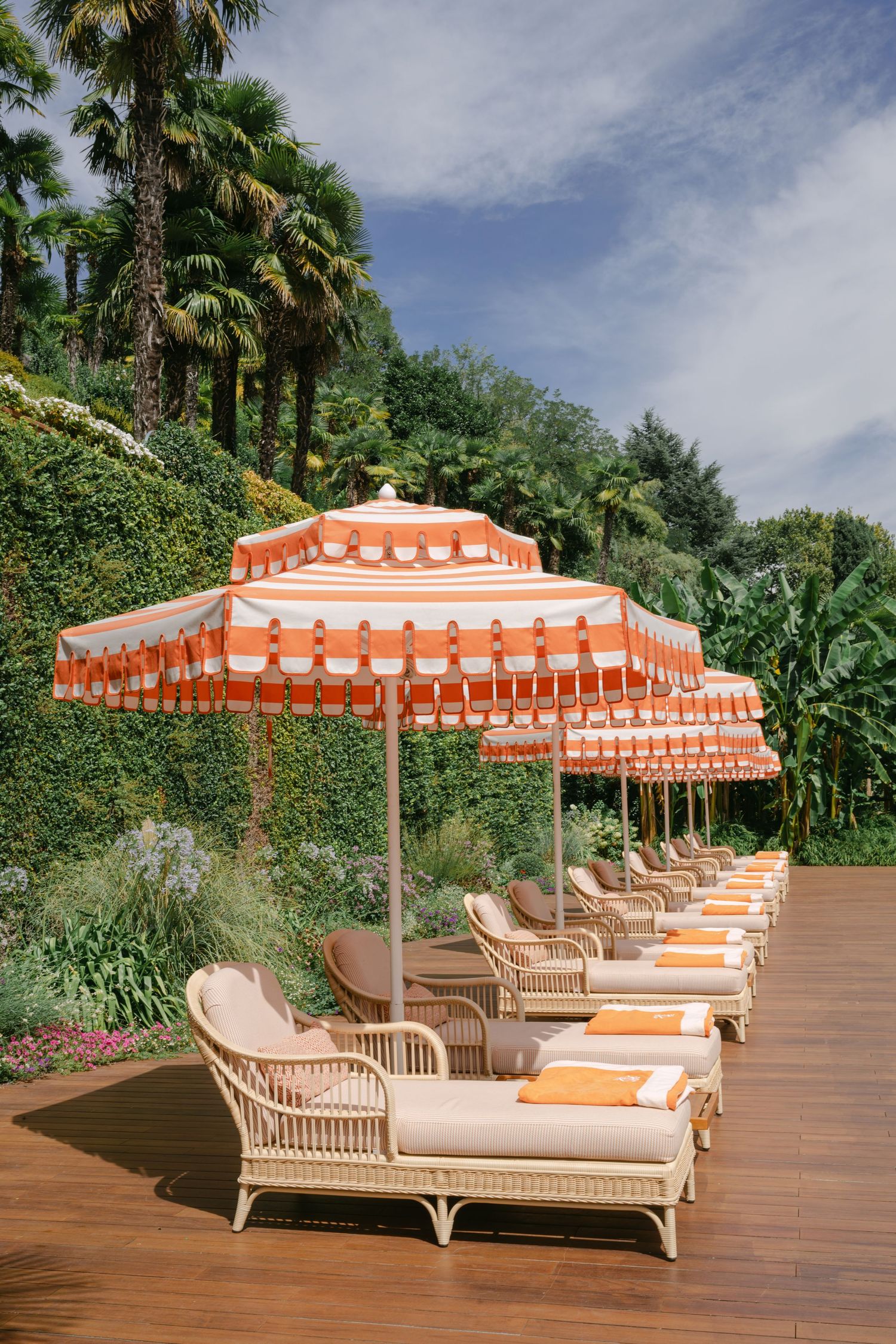Luxury rattan loungers with orange striped umbrellas on a wooden deck surrounded by lush tropical palms.