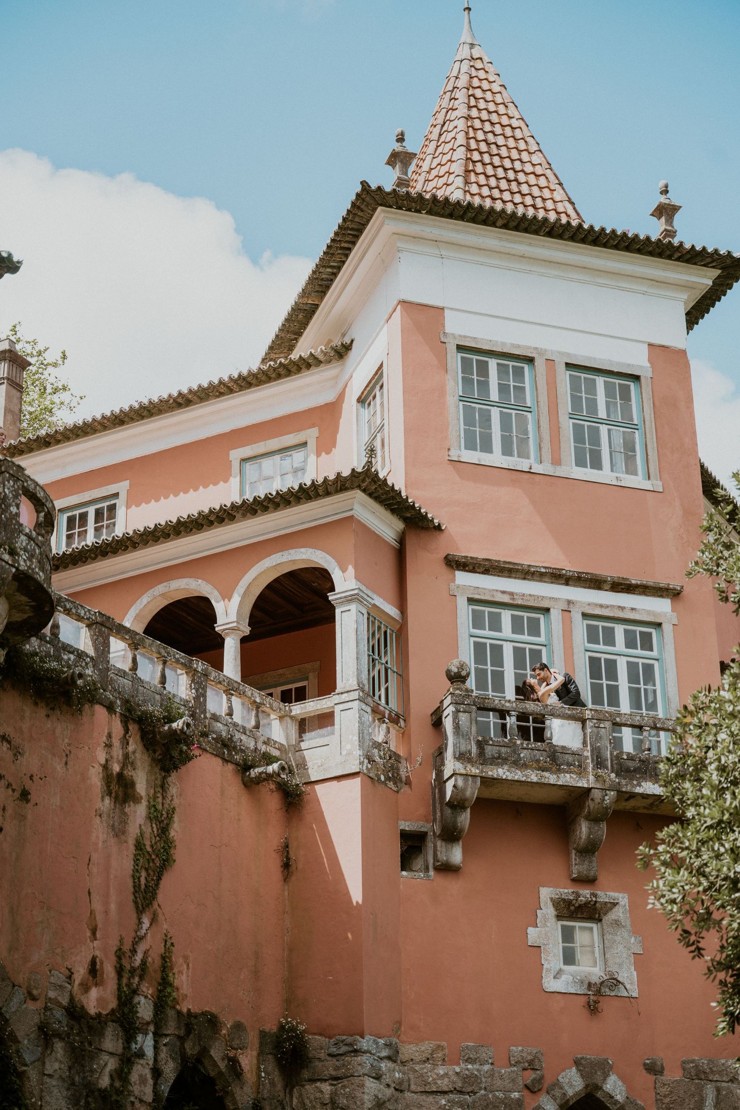 Couple on balcony of pink historic villa with terracotta roof and arched windows in Sintra, Portugal.