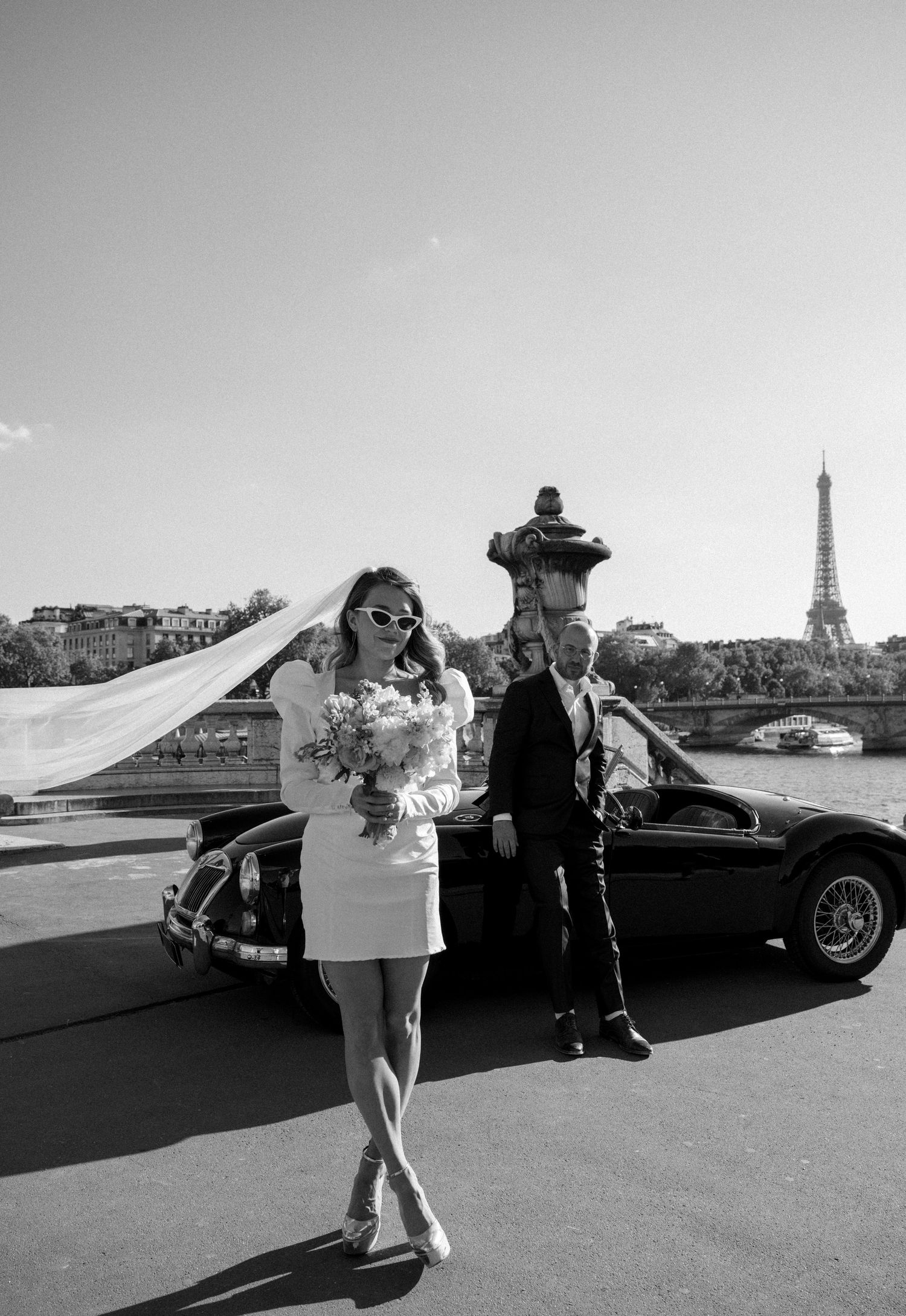 Bride in mini dress and veil poses with Eiffel Tower backdrop near vintage car in Paris.