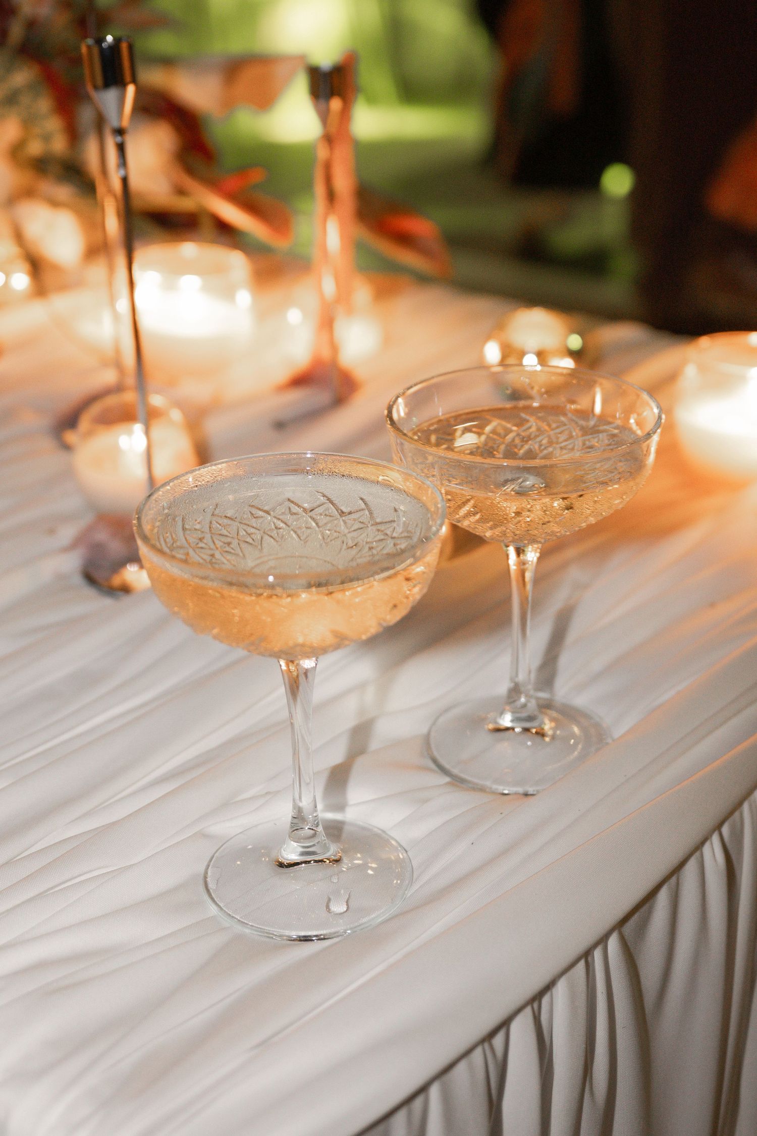 Two elegant crystal coupe glasses filled with champagne on a white draped table with candles at a wedding reception.