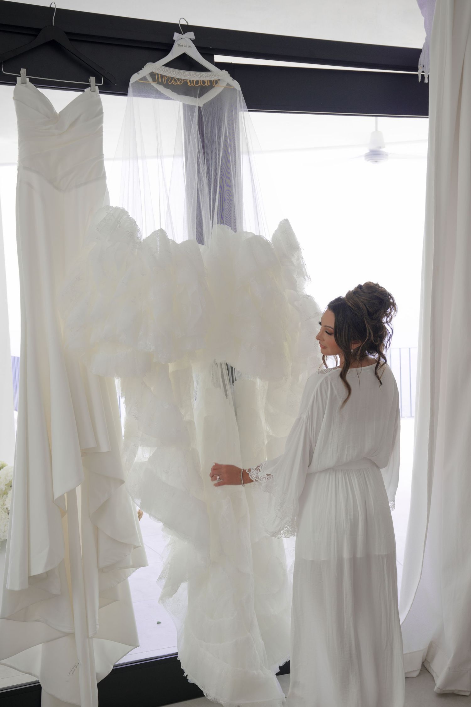 Bride in white robe admires her ruffled wedding gown hanging by a bright window before the ceremony.