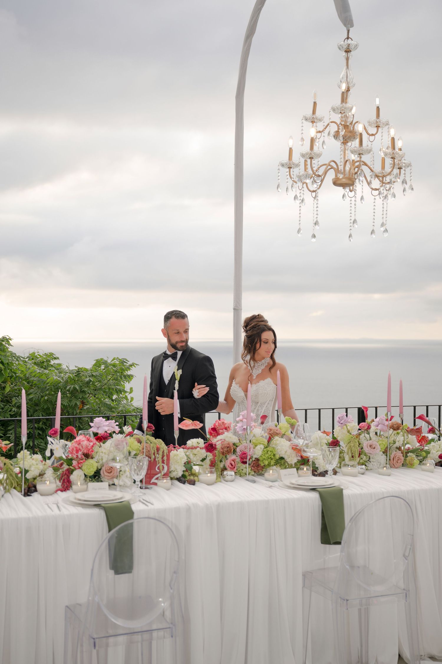 Bride and groom standing at elegantly decorated outdoor reception table with chandelier and ocean view.