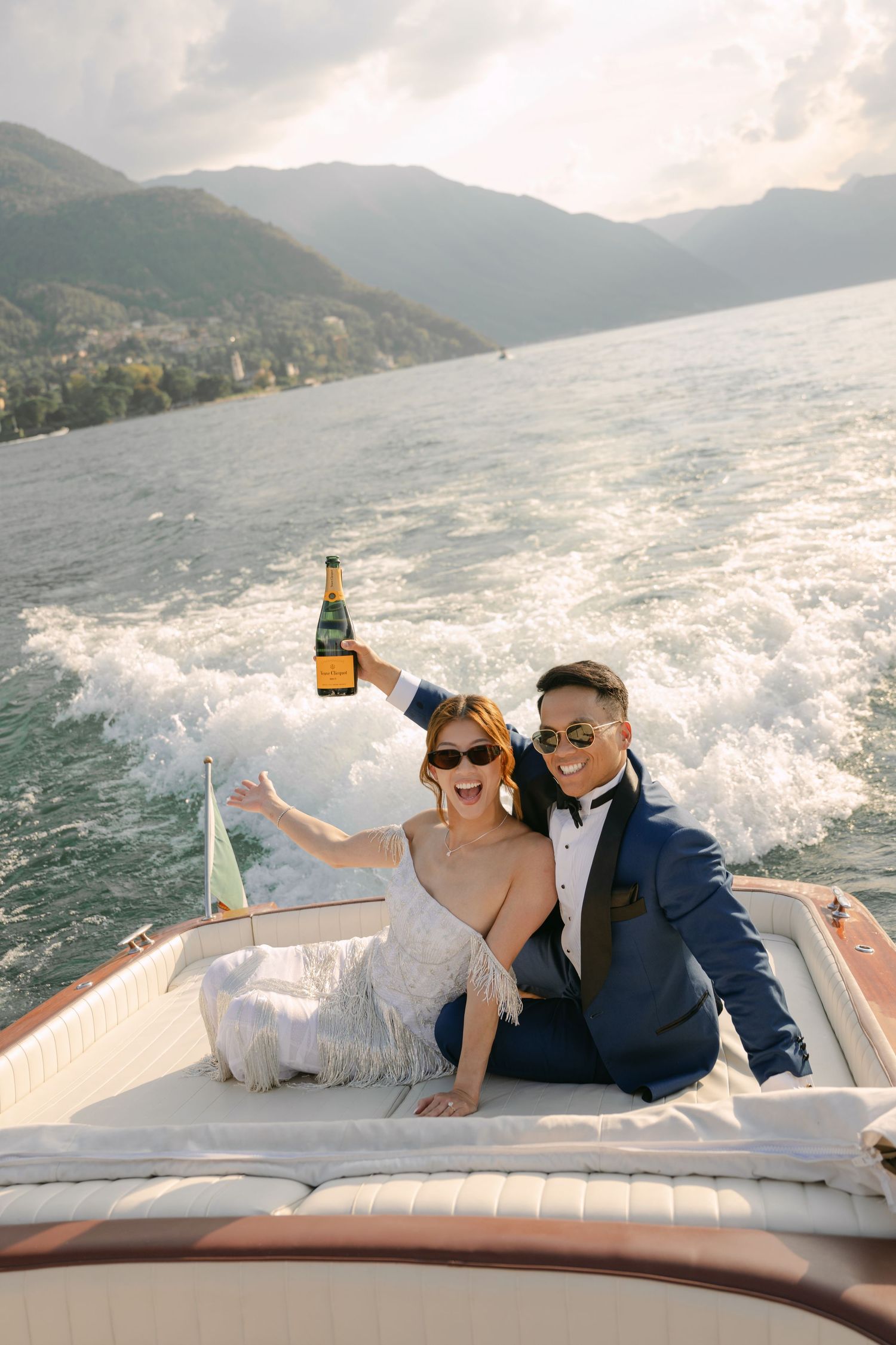 Couple celebrating on a luxury speedboat on Lake Como, holding champagne, dressed in wedding attire with mountains behind.
