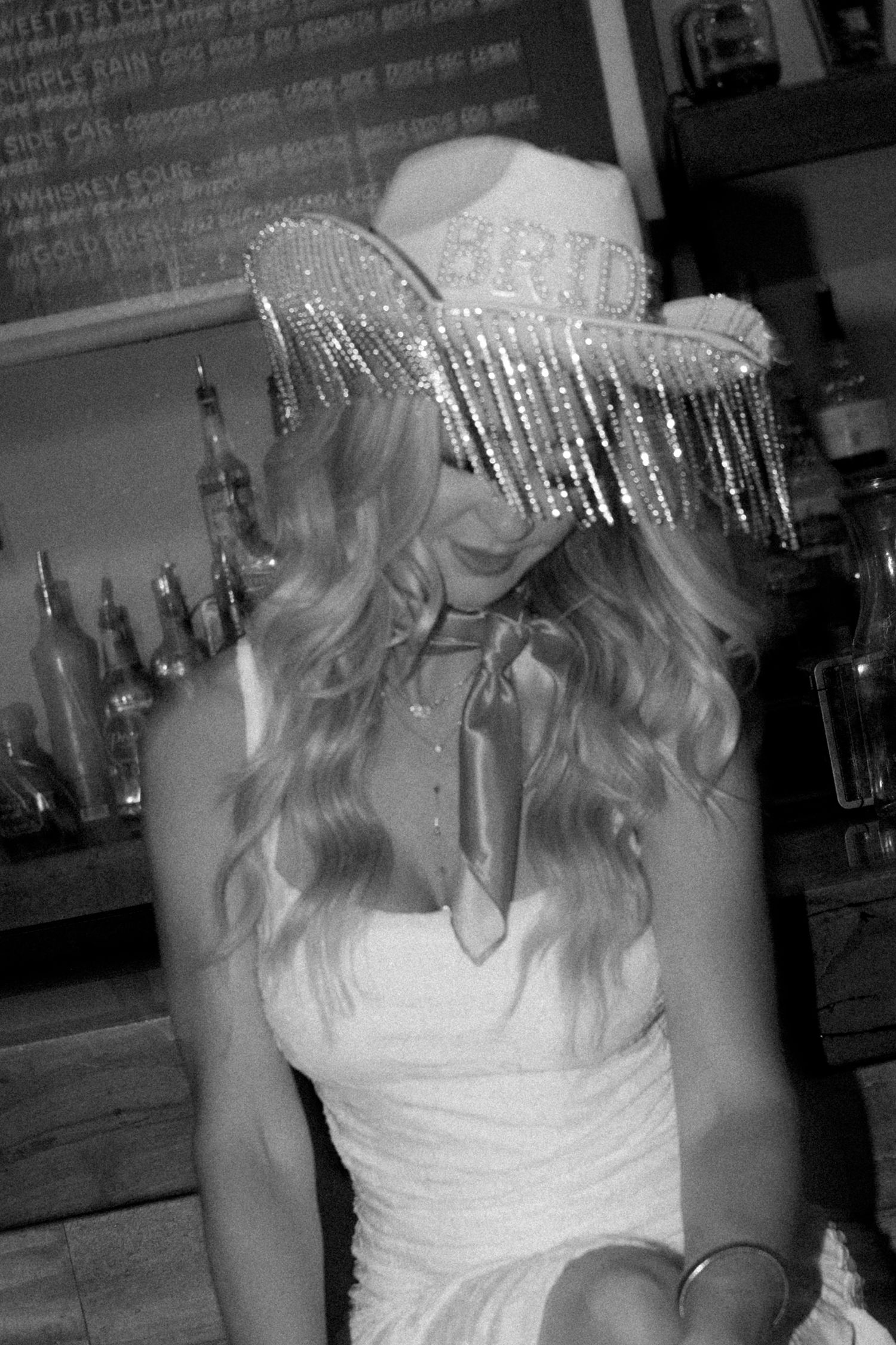 Black and white photo of a blonde woman wearing a rhinestone BRIDE cowboy hat and white dress at a bar.
