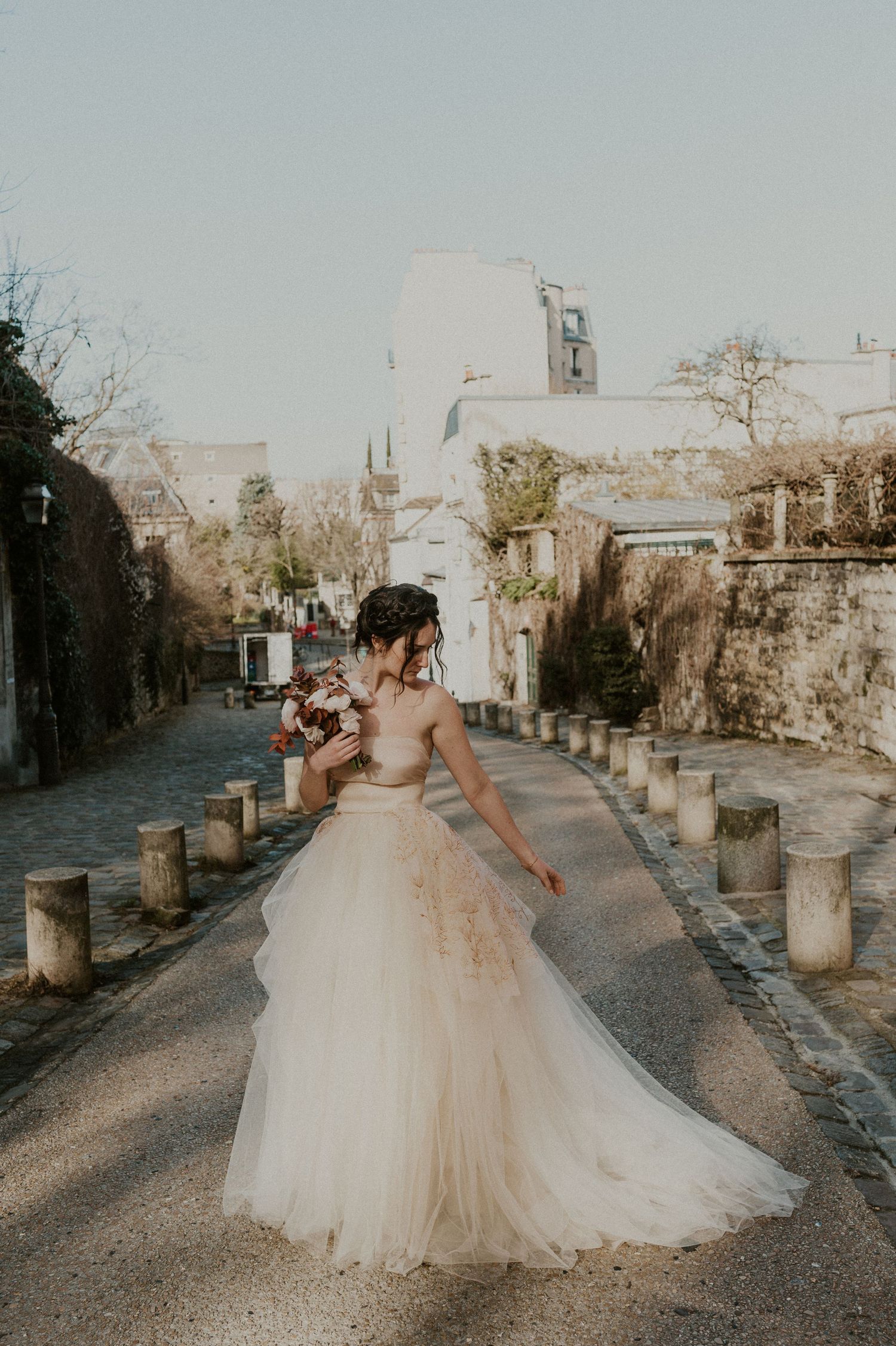 A bride in a flowy tulle wedding gown poses on a cobblestone path lined with stone pillars in a European setting.