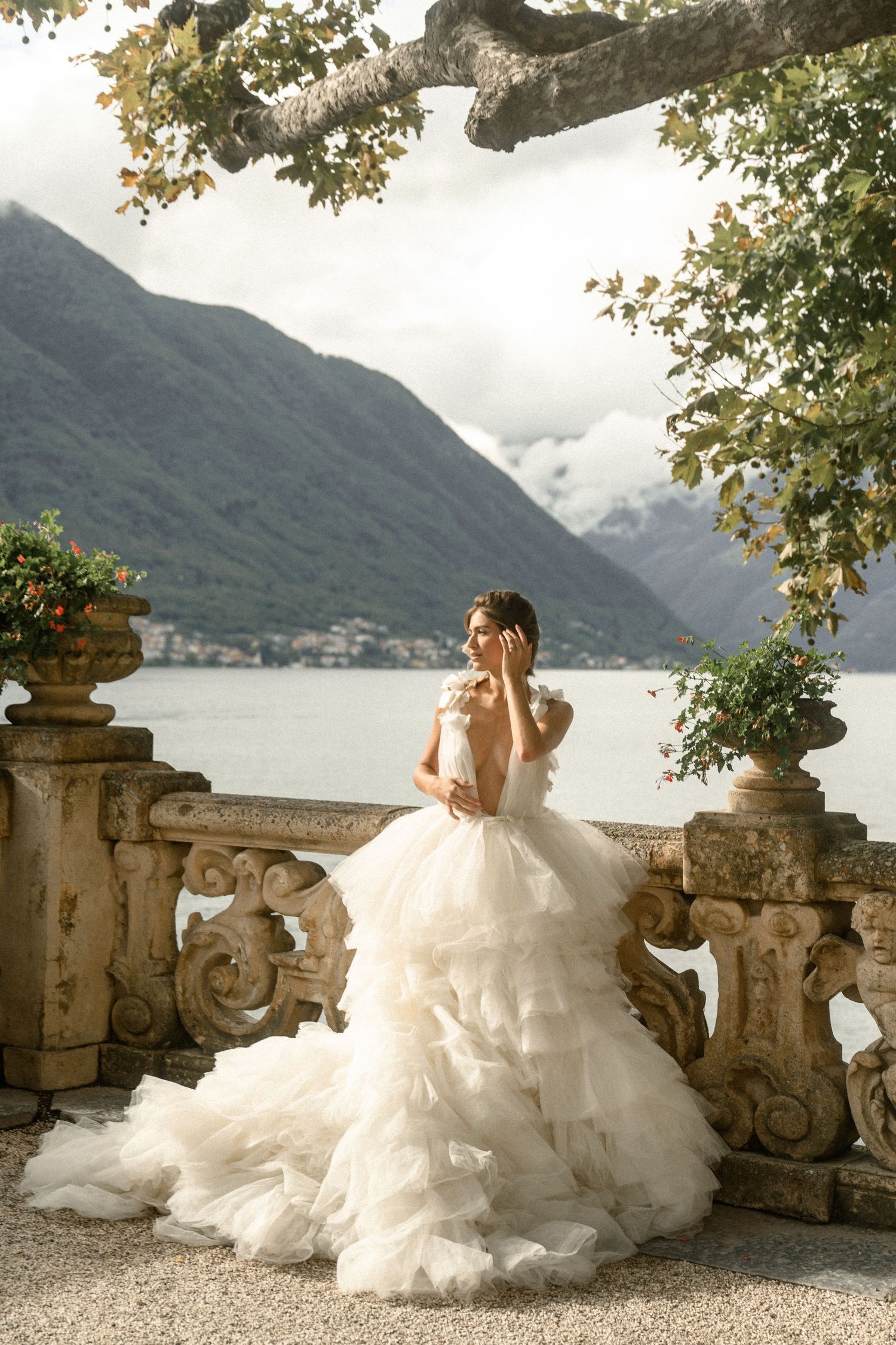 A bride in a layered tulle gown poses on a lakeside terrace with mountain views.