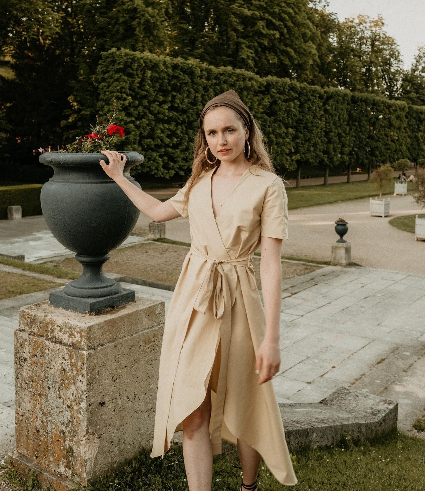 A beige wrap dress worn in an elegant garden setting with a decorative urn and manicured hedges.