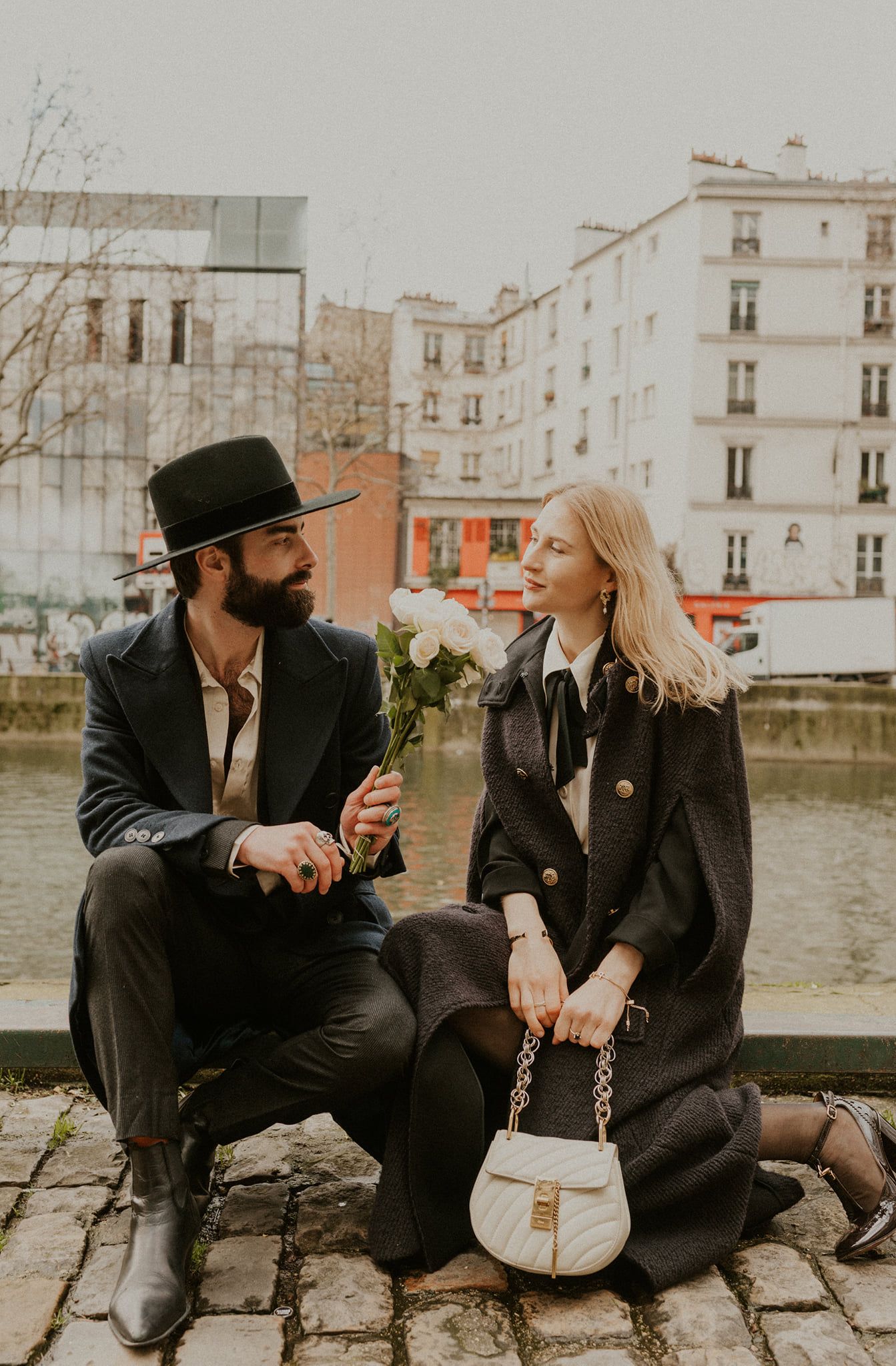 A stylish couple shares a moment along a Parisian canal on a winter day.