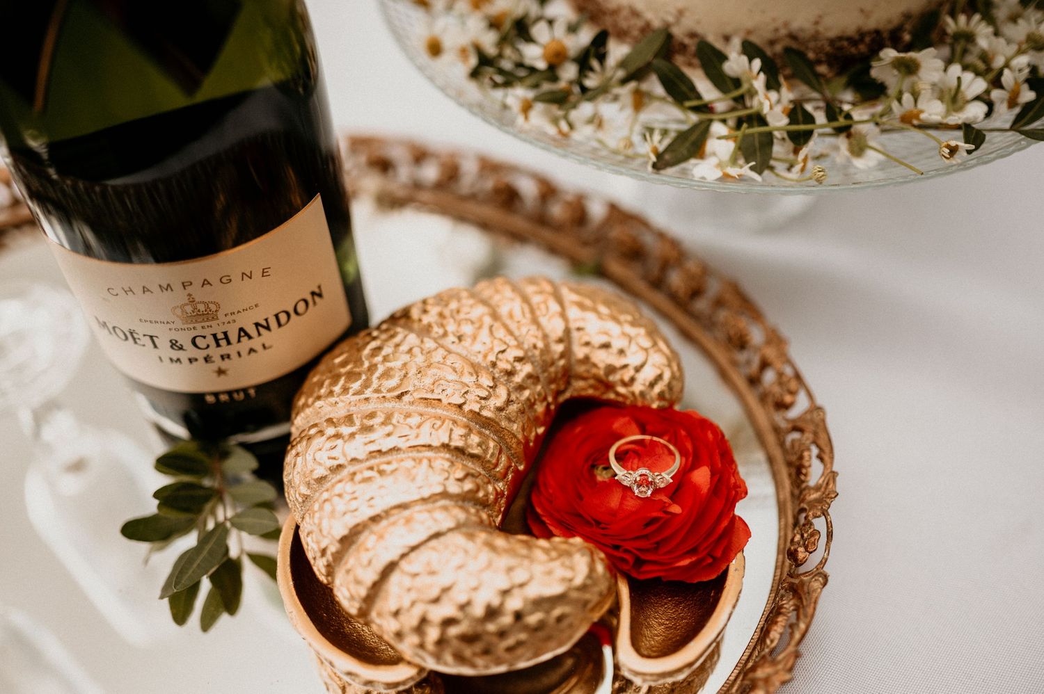 A champagne bottle and gold dessert plate with a red ring box.