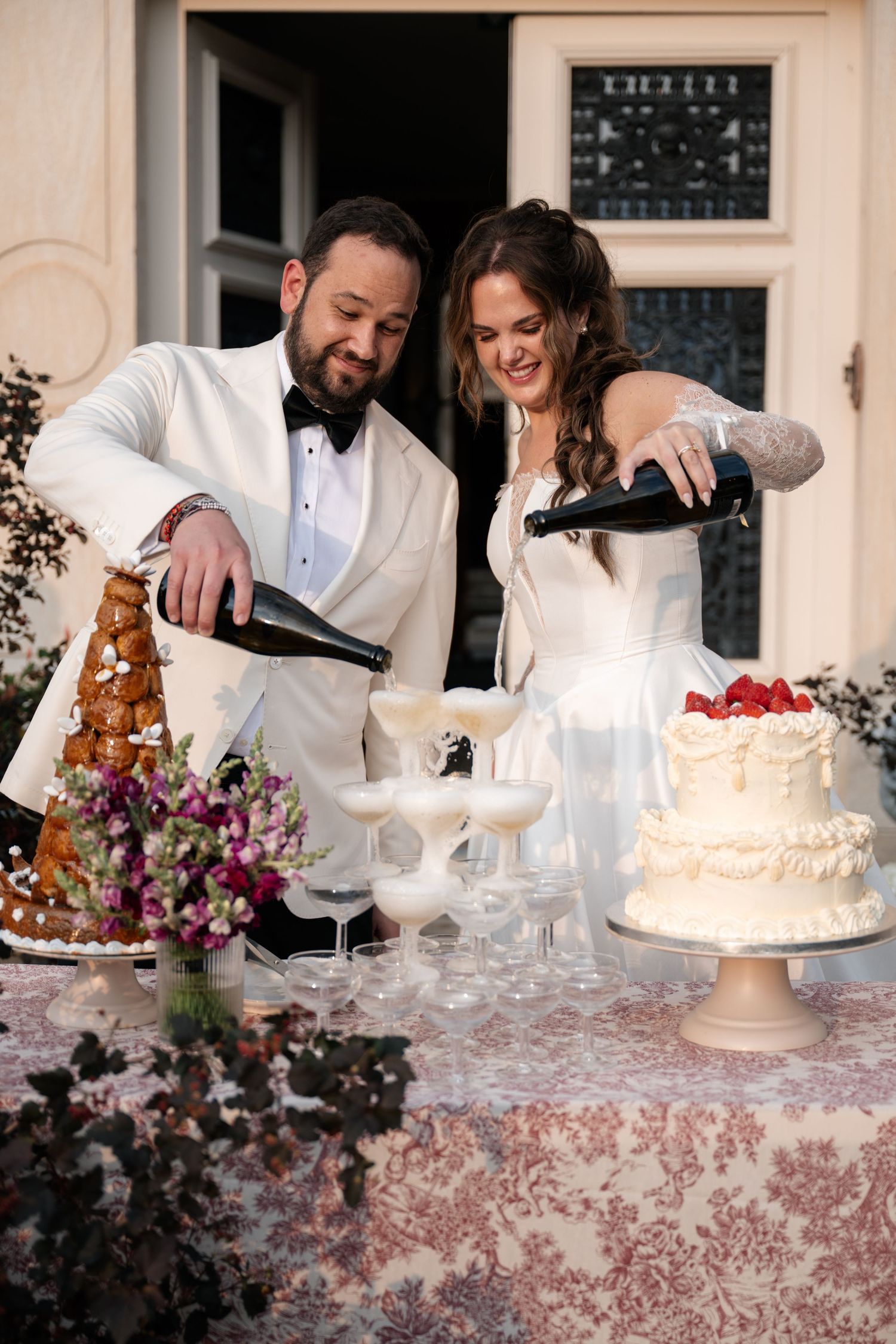 Wedding couple cutting and serving elegant white tiered cakes at an ornate dessert table.