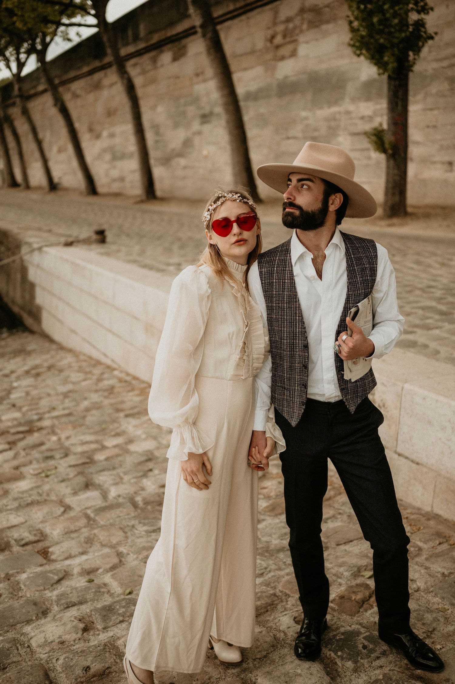 A sepia-toned portrait of a couple in vintage clothing with red sunglasses against a stone wall.