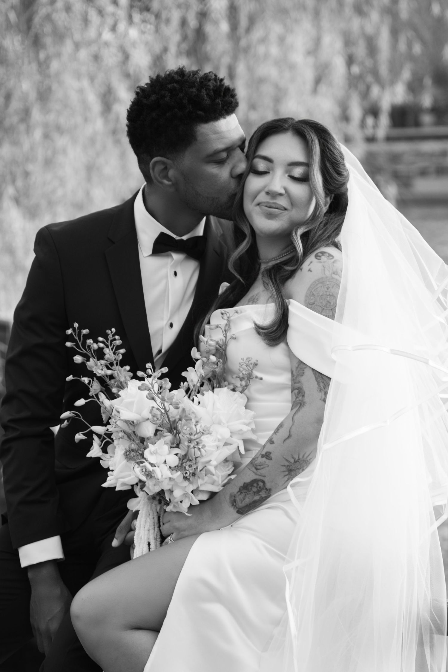 Black and white portrait of groom kissing bride holding blue and white bouquet under willow tree.
