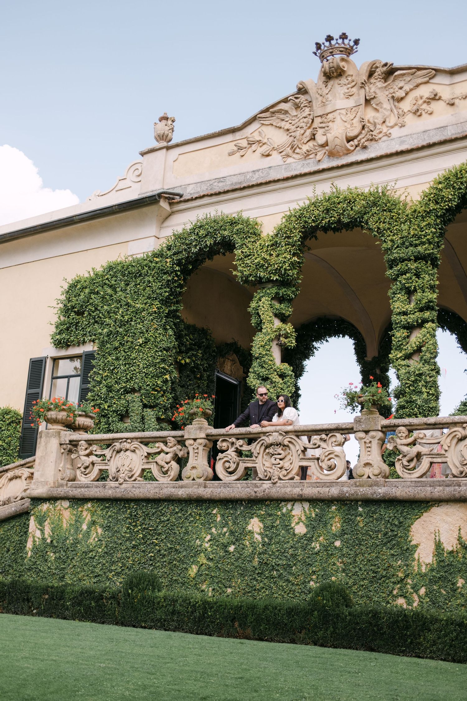 Couple stands on ornate stone balcony of ivy-covered Italian villa with decorative arches and coat of arms.