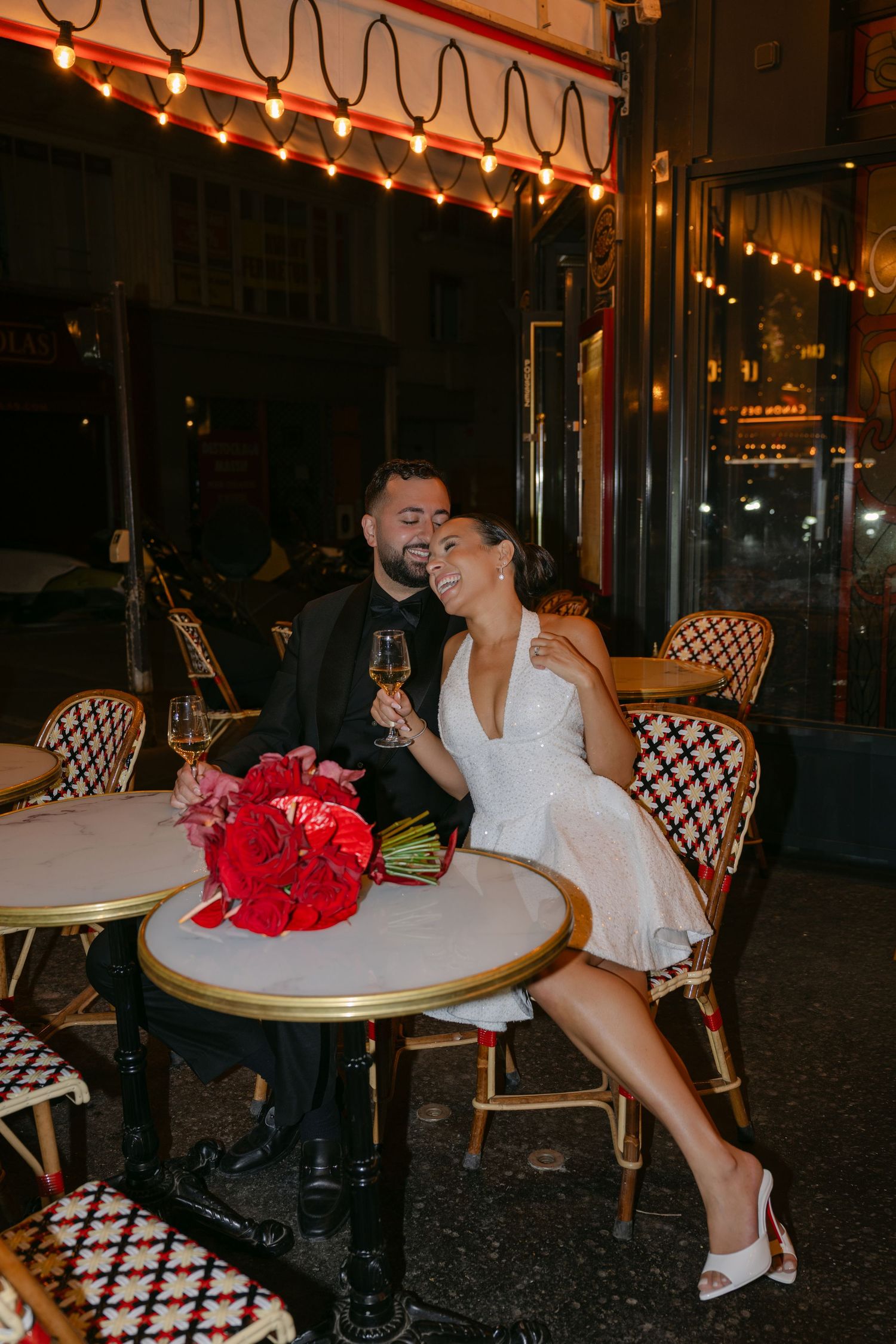 Couple celebrating at a Parisian cafe at night, woman in white dress holding wine glass with red peonies on table.