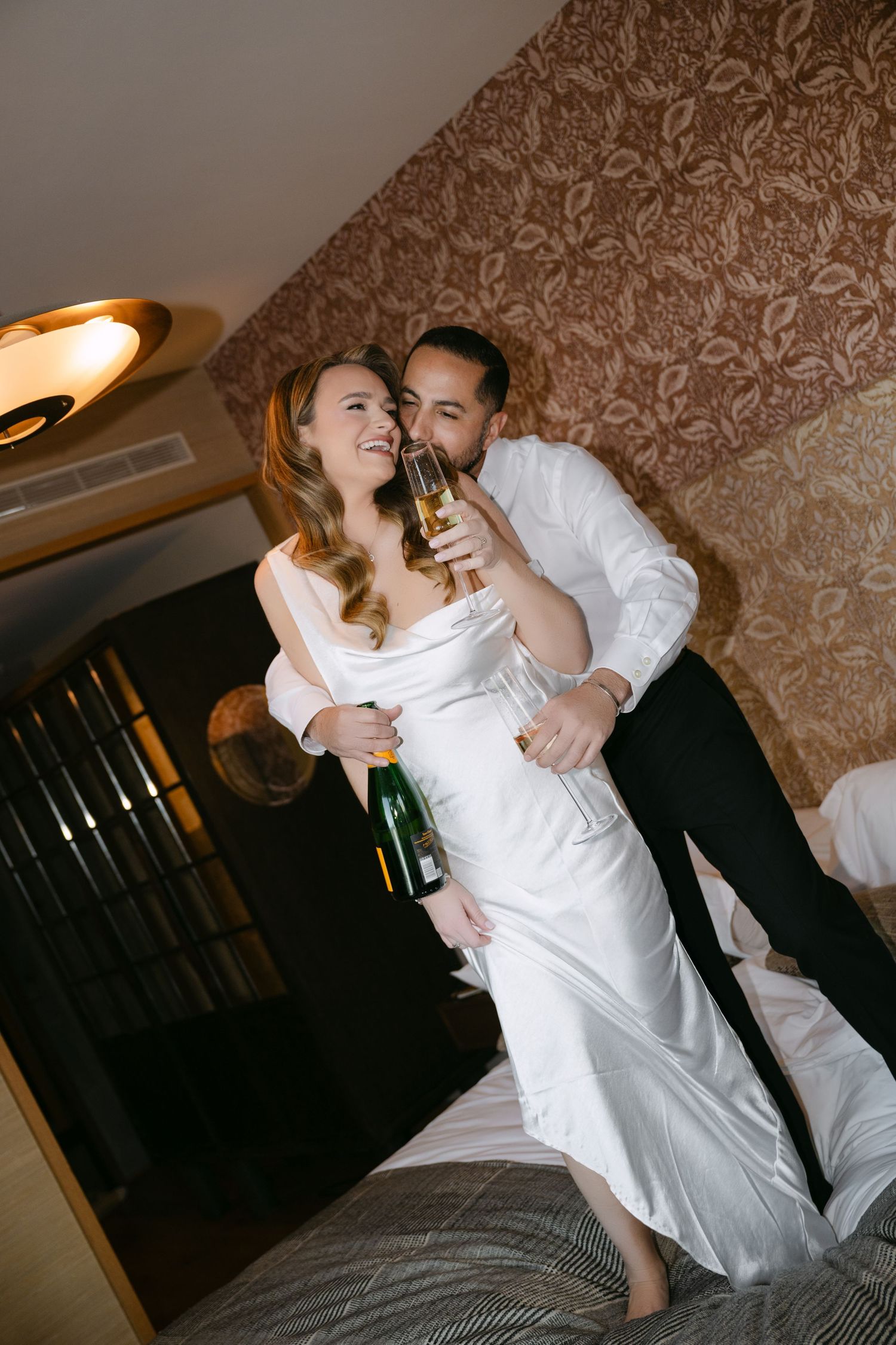 Bride and groom celebrating wedding day with champagne in elegant hotel room, sharing a romantic toast together.