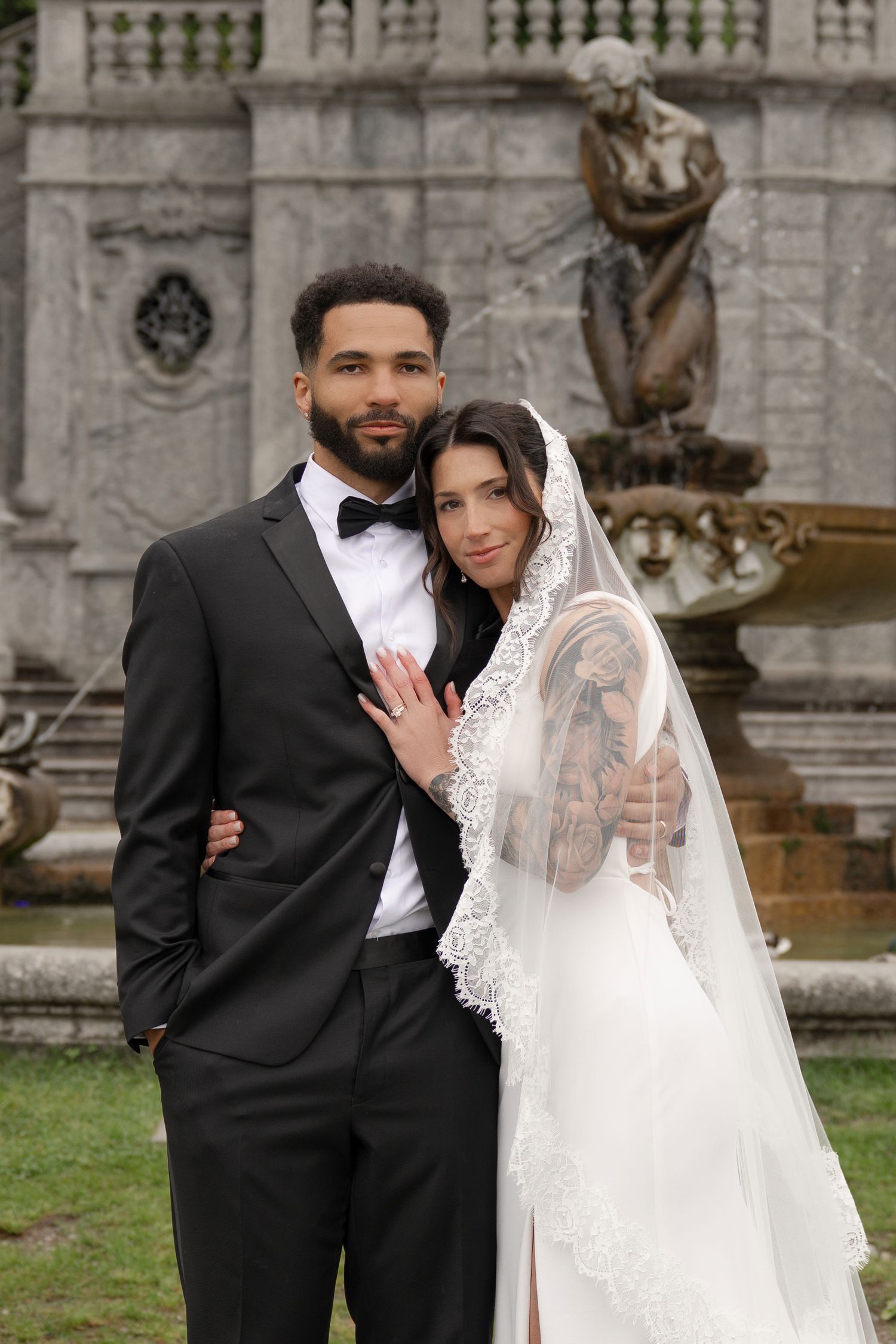 Interracial wedding couple poses before ornate stone fountain, bride in lace veil and groom in black tuxedo.
