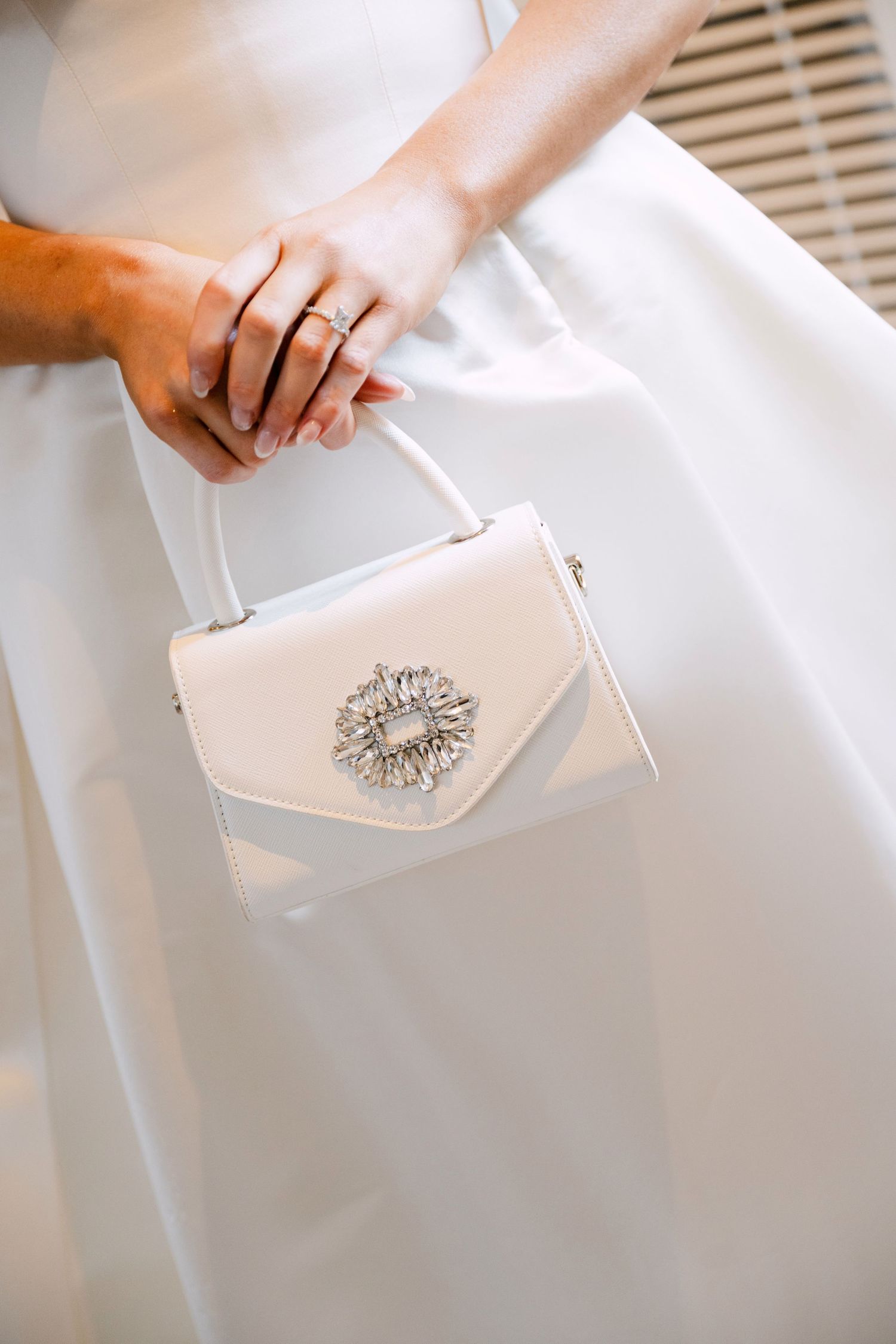 Bride holding an elegant white satin mini handbag with crystal brooch detail while wearing a white wedding gown.