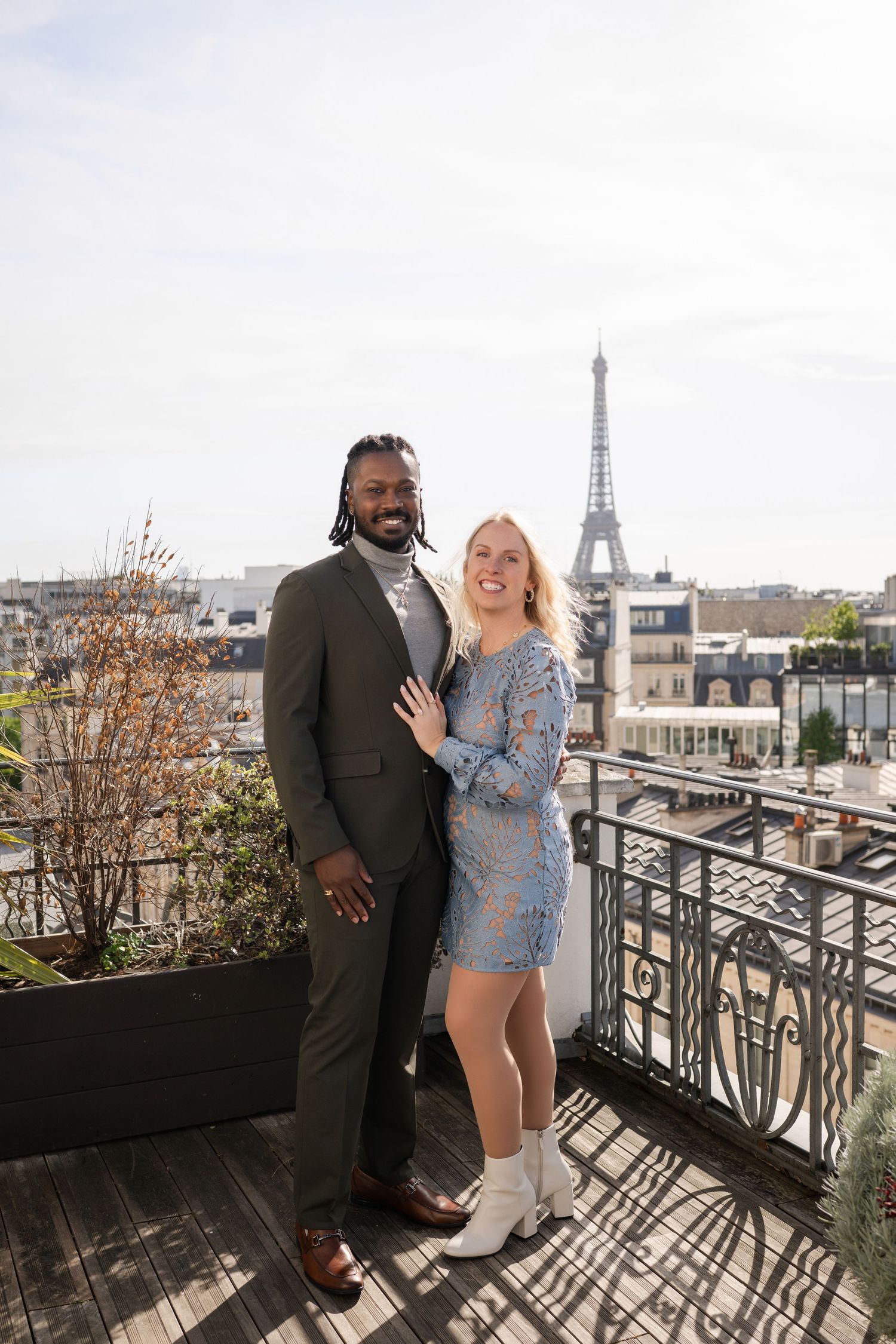 Couple posing on Paris rooftop terrace with Eiffel Tower backdrop, woman in blue lace dress, man in olive suit.