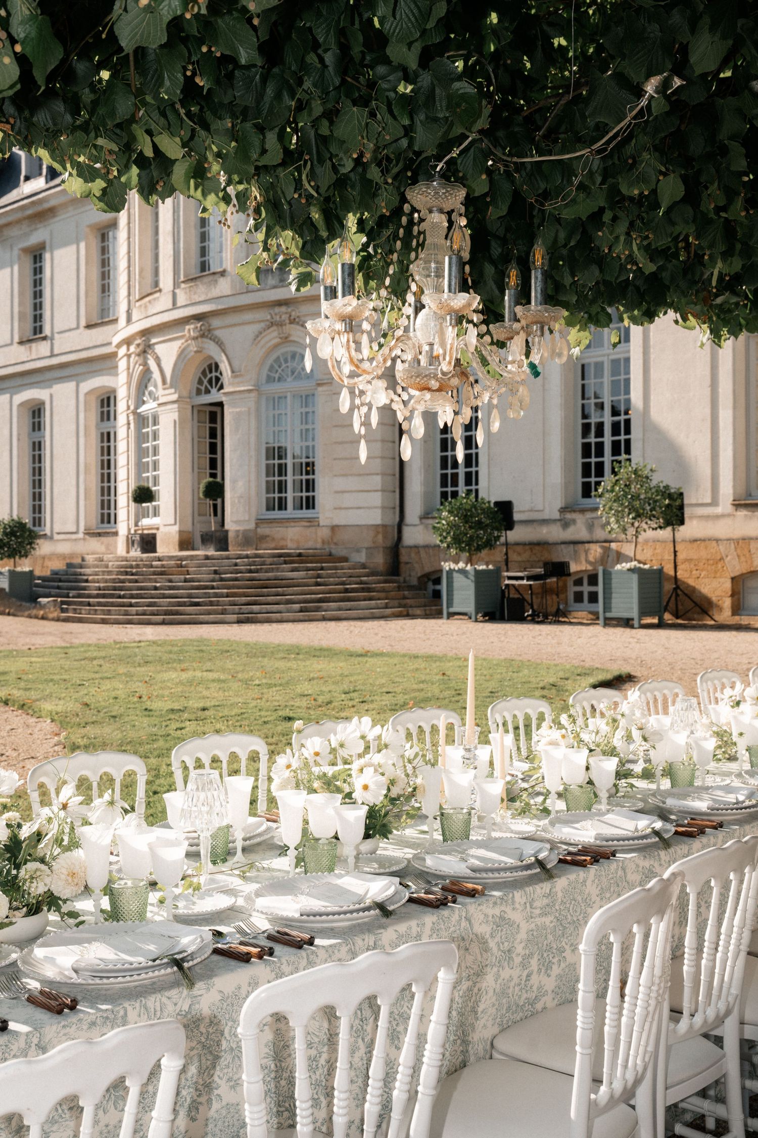 Elegant outdoor wedding reception table with chandelier beneath tree at French chateau.