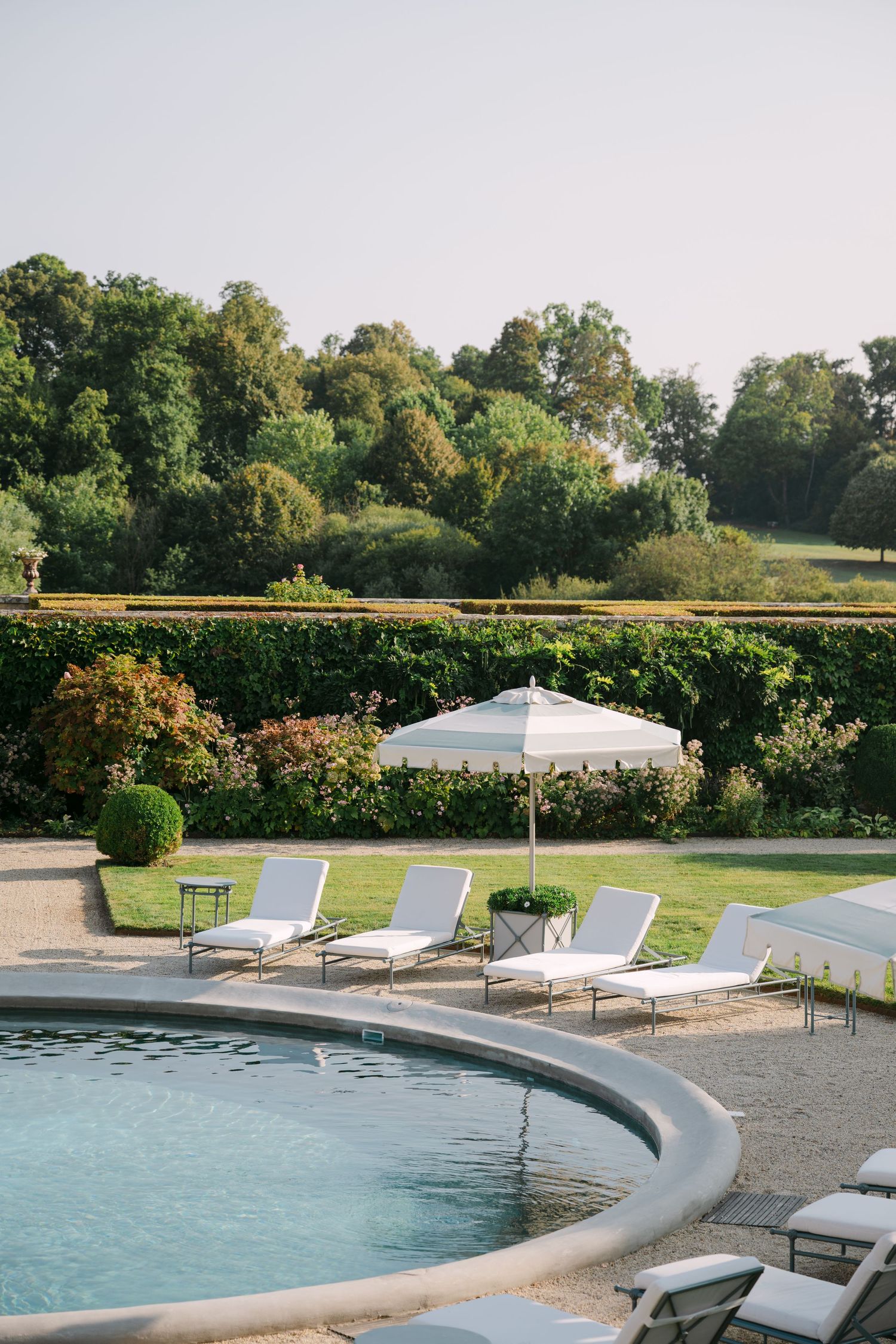 Curved luxury pool edge with white sun loungers and a striped umbrella set against lush green hedged gardens.