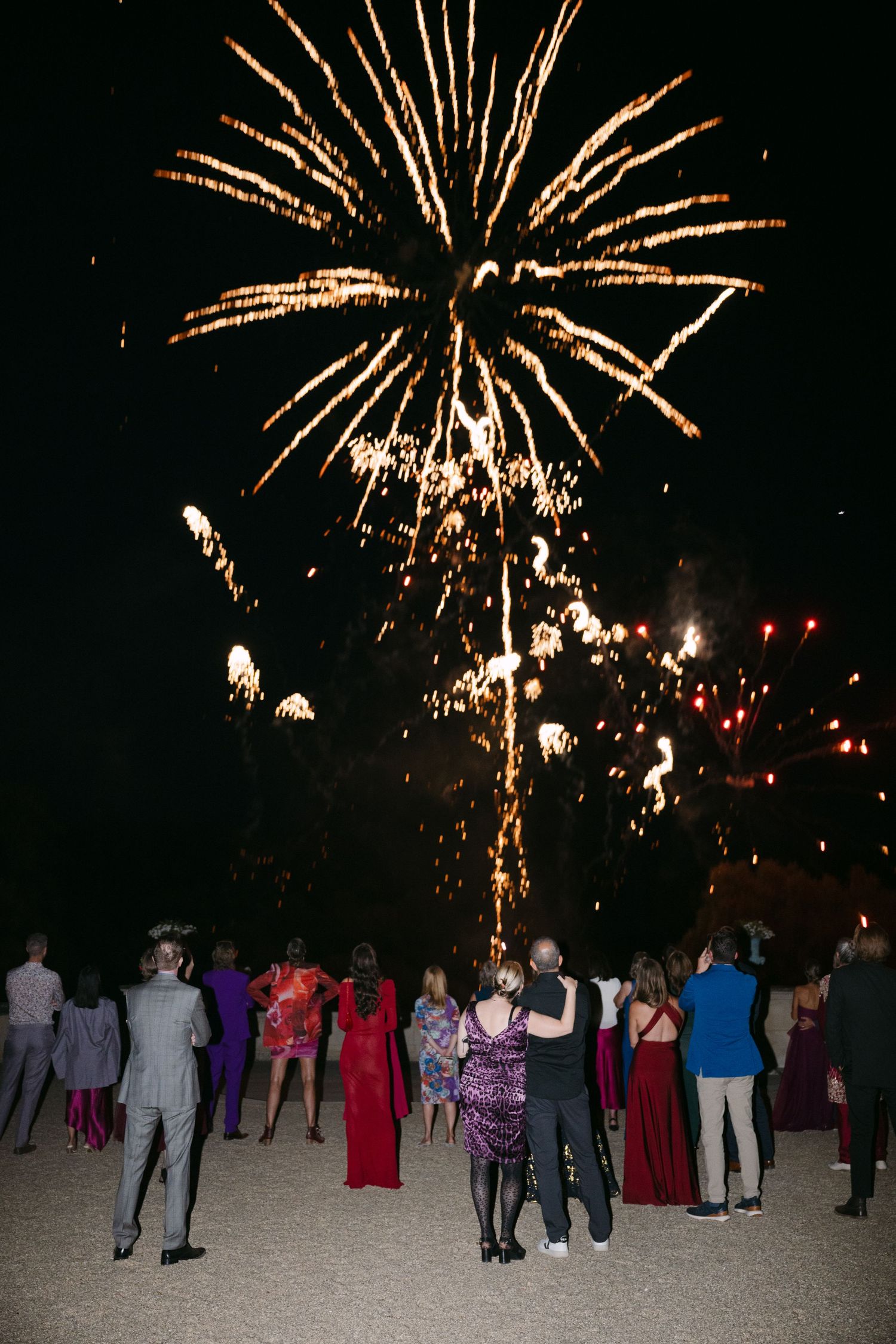Guests in formal attire watch a stunning golden fireworks display light up the night sky at an outdoor celebration.