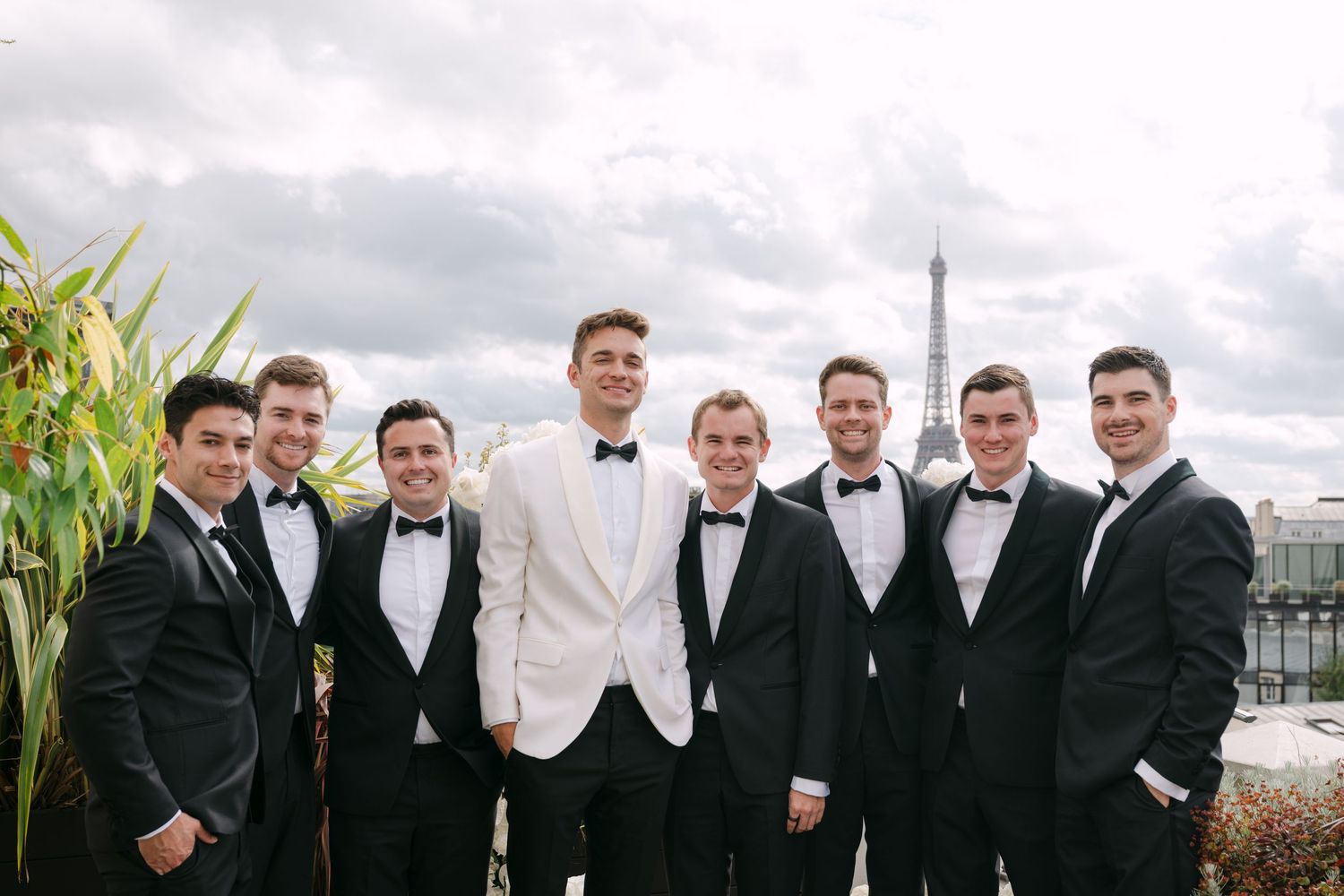 Groom in white tuxedo poses with seven groomsmen in black tuxedos on Paris rooftop terrace.