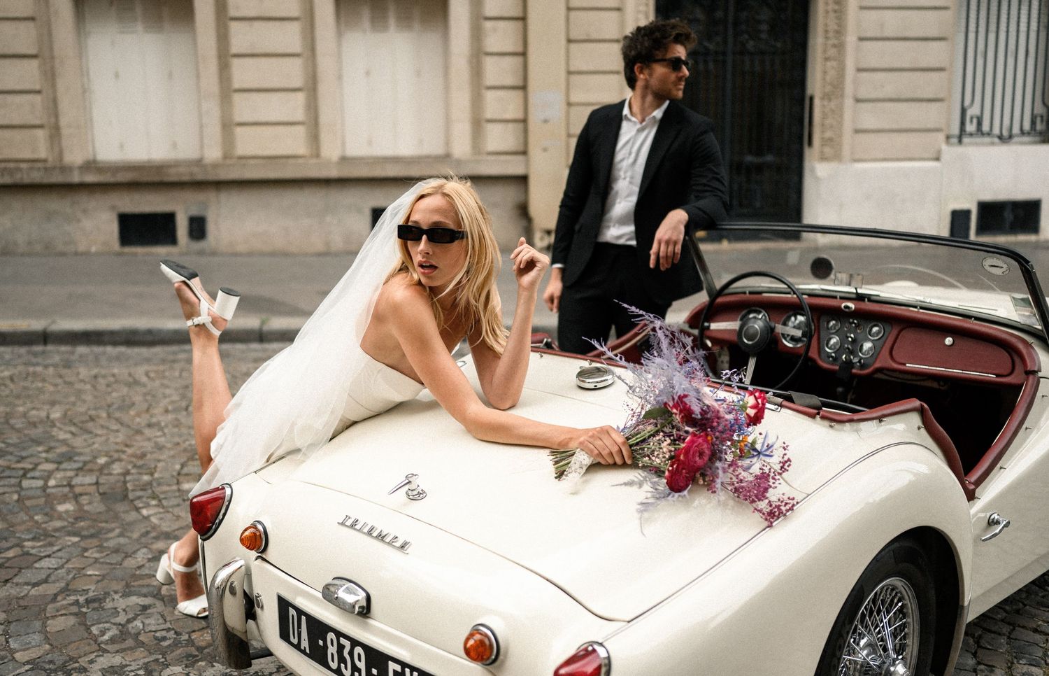 Stylish bride in sunglasses poses on vintage white Triumph car while groom stands behind on Paris cobblestone street.