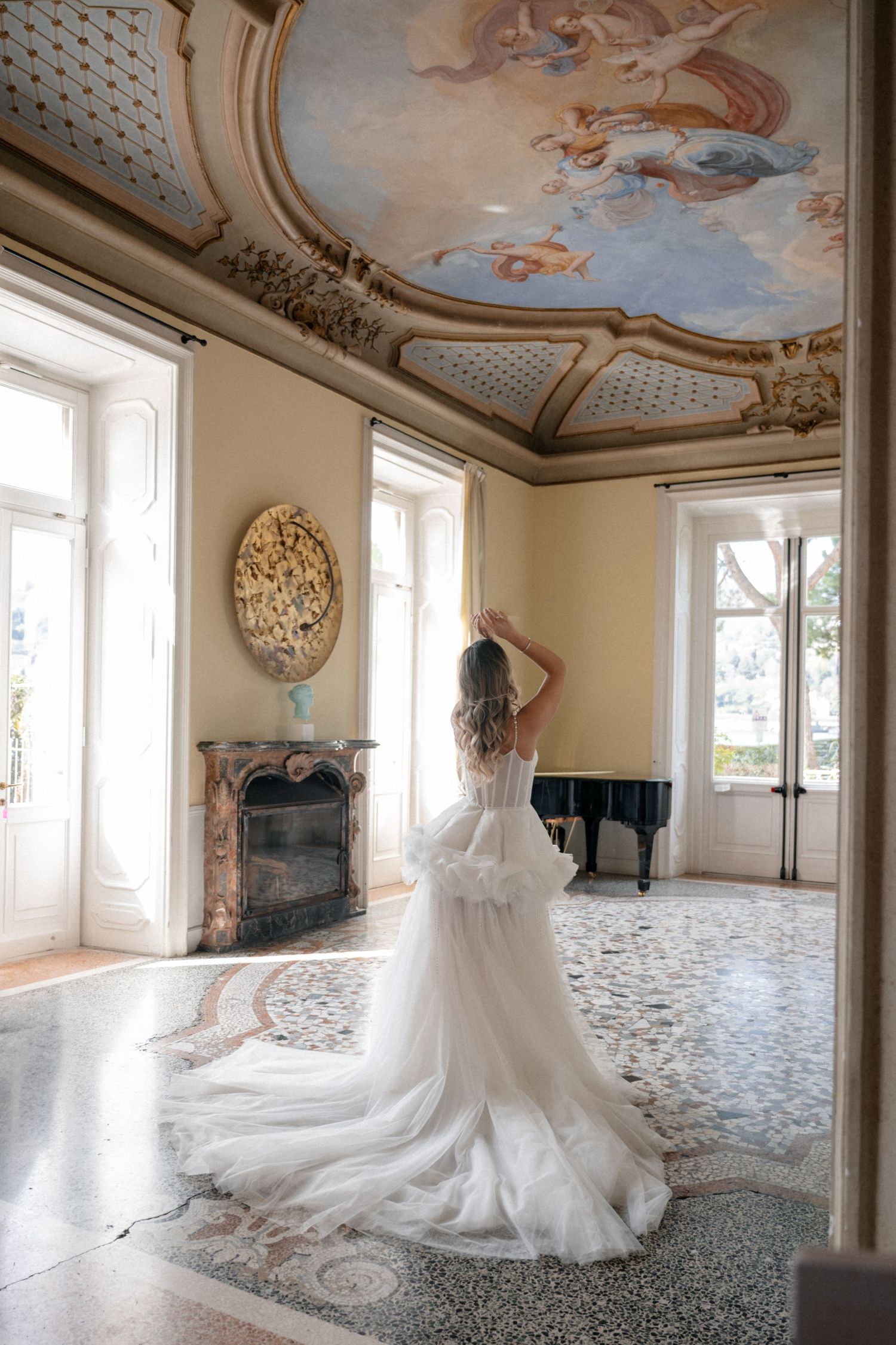Bride in white ruffled gown stands in ornate villa room with baroque ceiling fresco and grand piano.