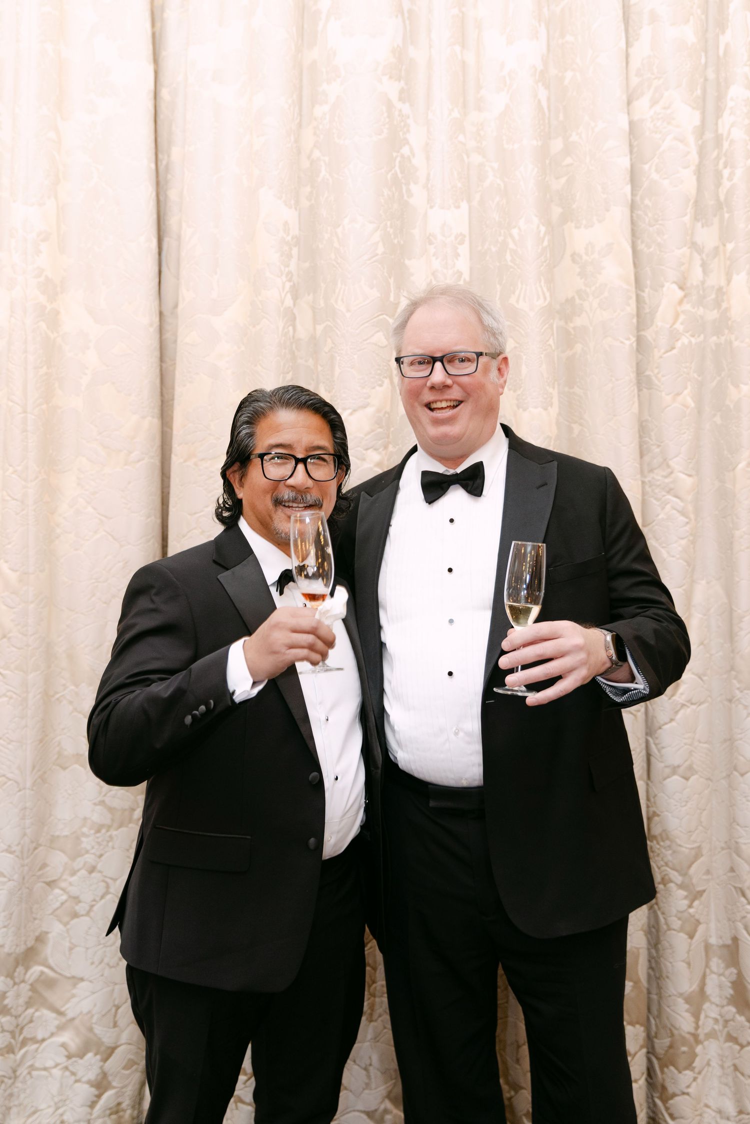 Two men in black tuxedos toasting champagne glasses at elegant wedding reception photo booth.