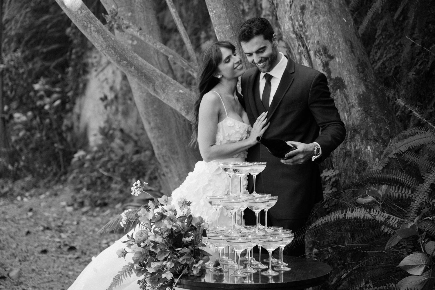 Black and white photo of a bride and groom sharing a romantic moment beside a champagne tower outdoors.