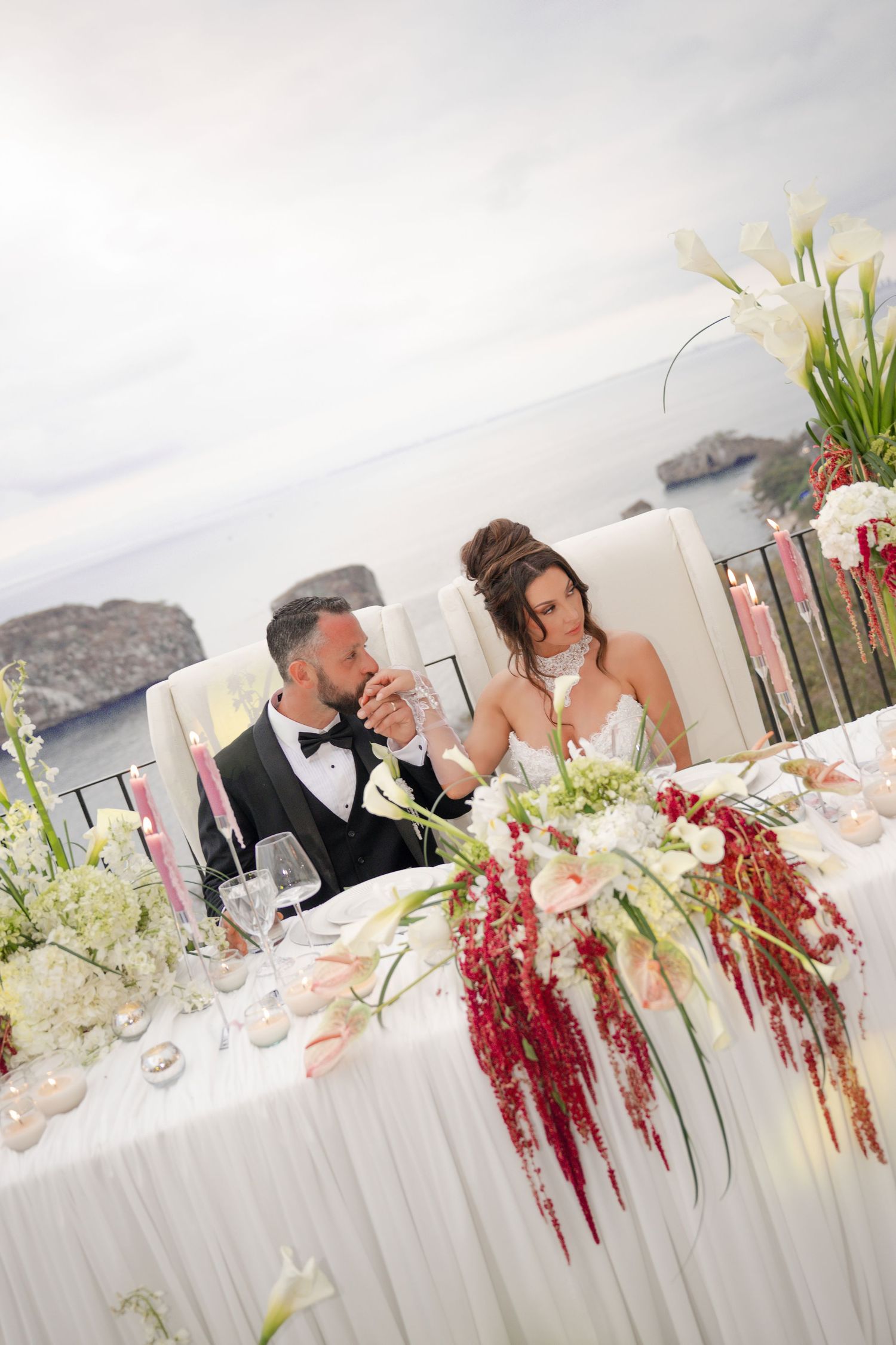 Bride and groom share a toast at their elegant outdoor wedding reception table adorned with white and red floral arrangements.