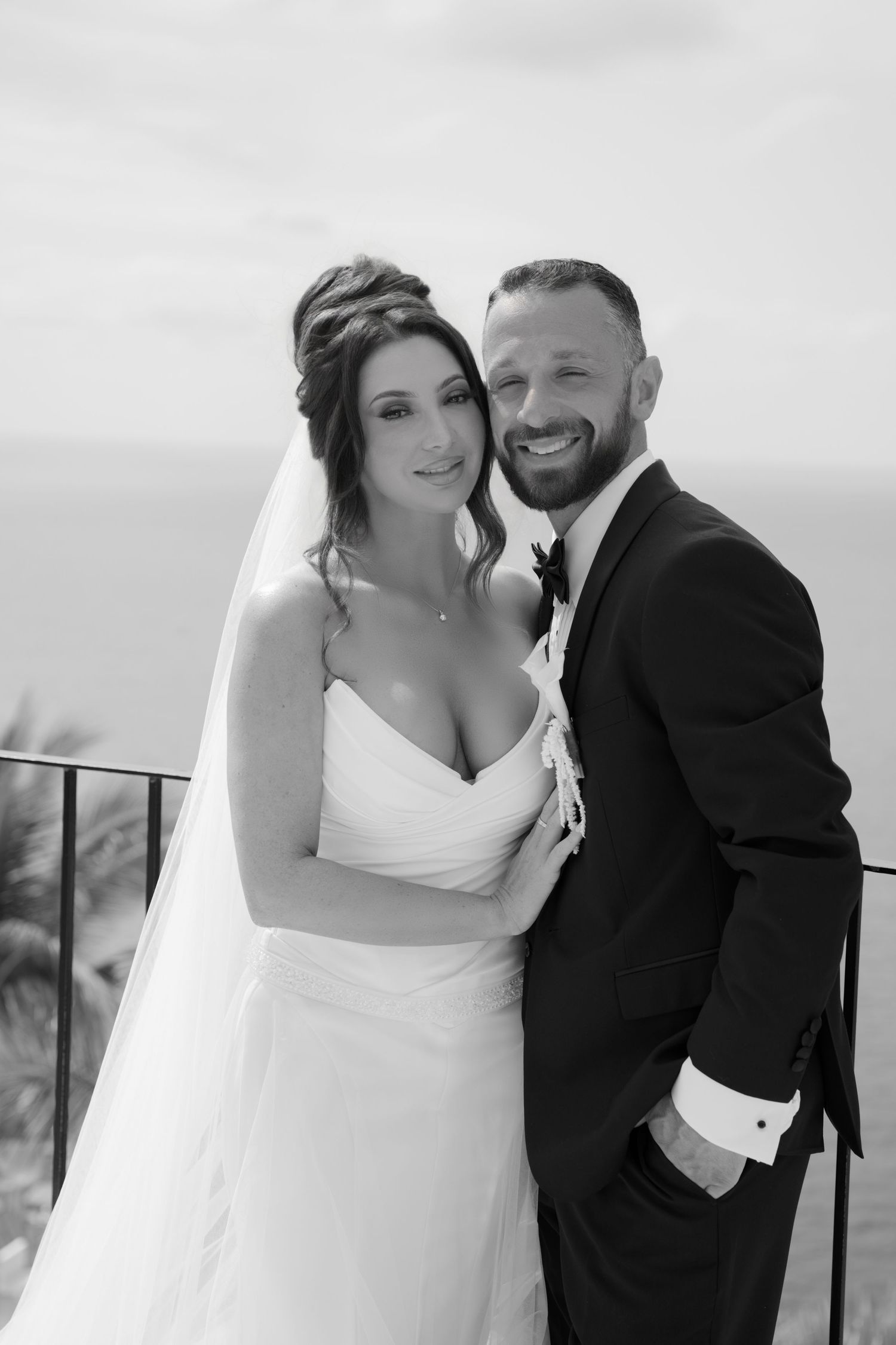 Black and white wedding portrait of smiling bride in white gown and groom in tuxedo posing together outdoors.