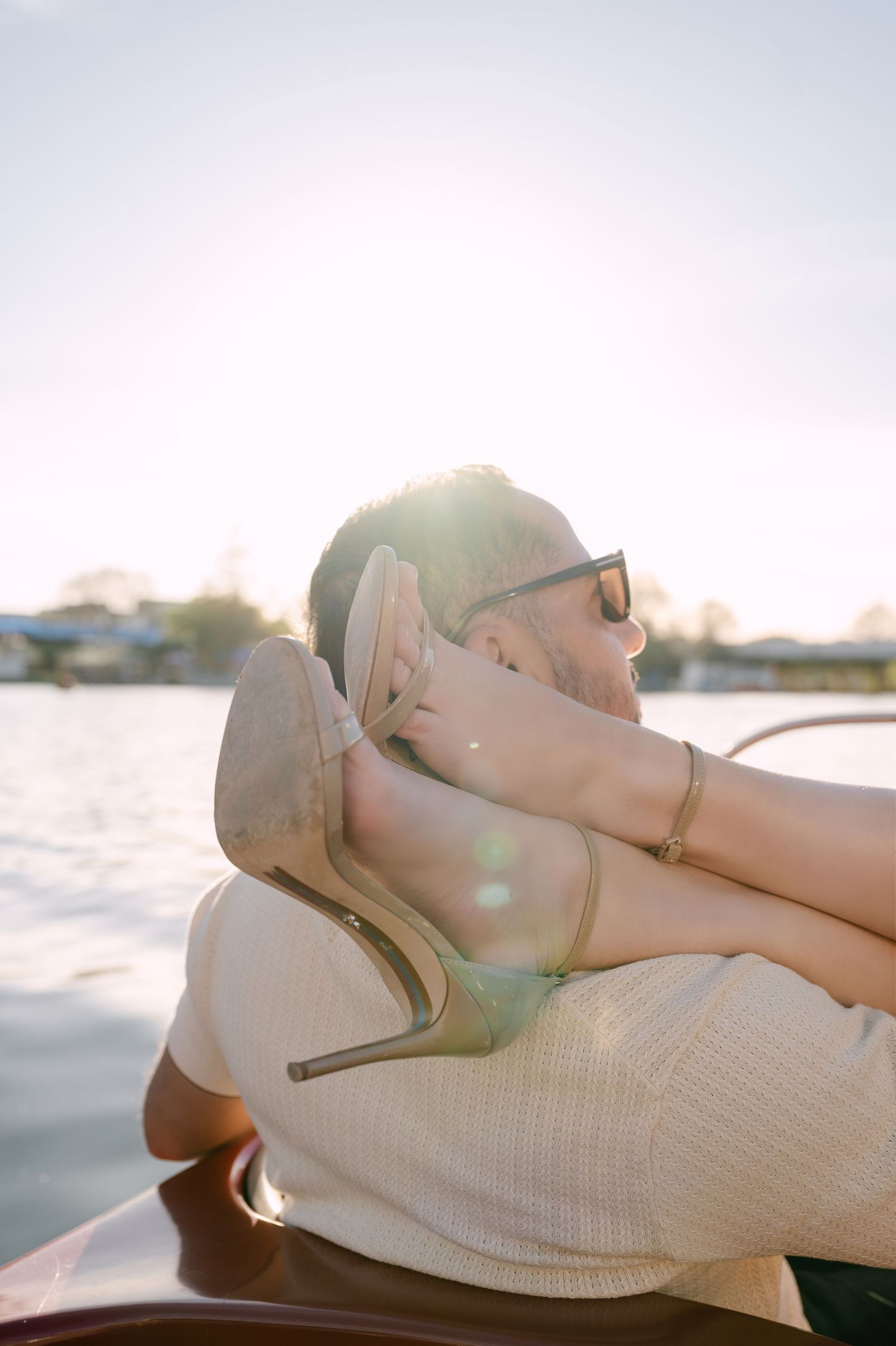 Couple sharing a romantic moment on a boat at sunset, woman's heeled shoe visible as sun flares over water.