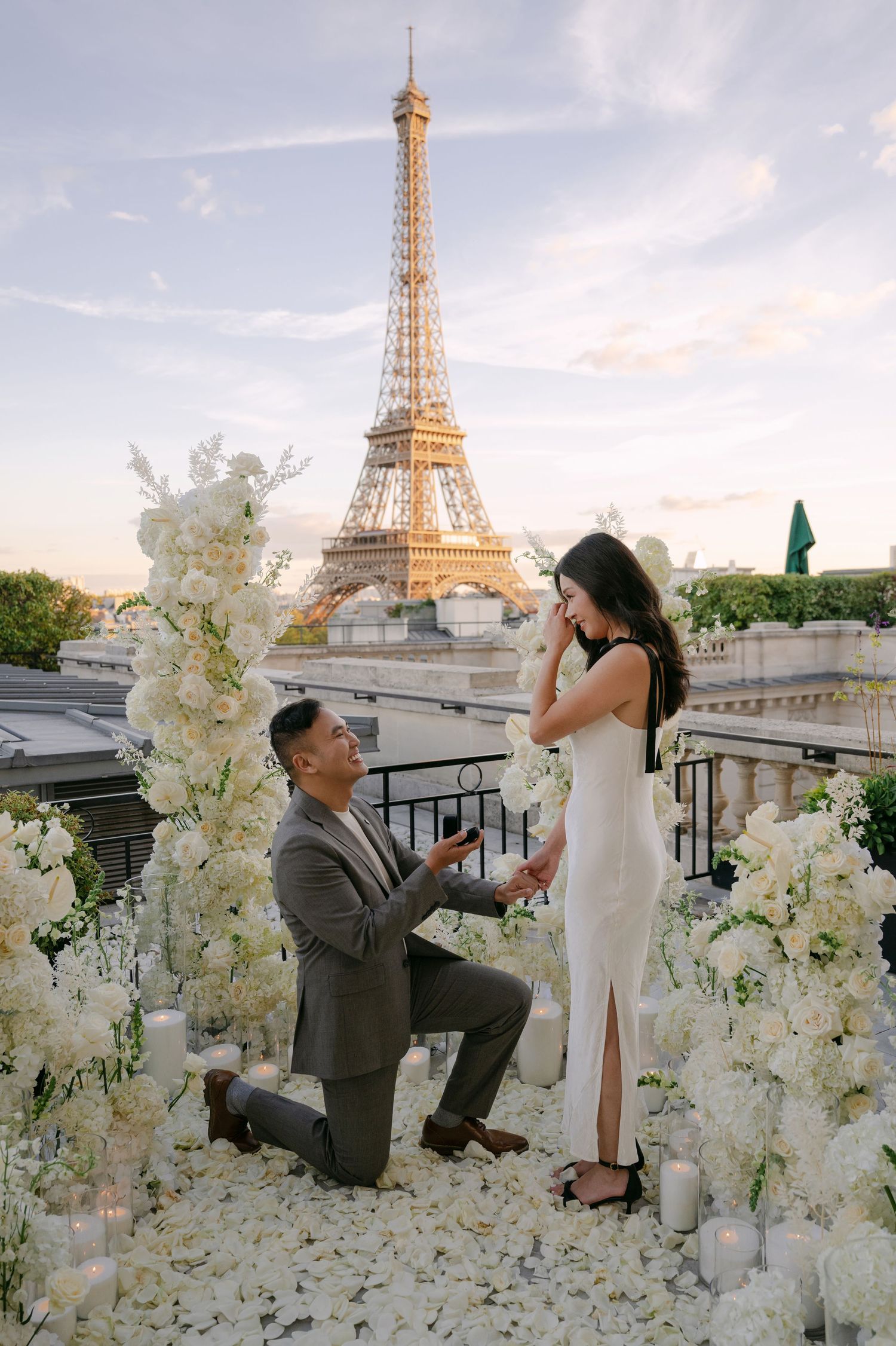 Man kneels with a ring box as a woman in a white dress covers her face on a Paris rooftop with the Eiffel Tower behind.