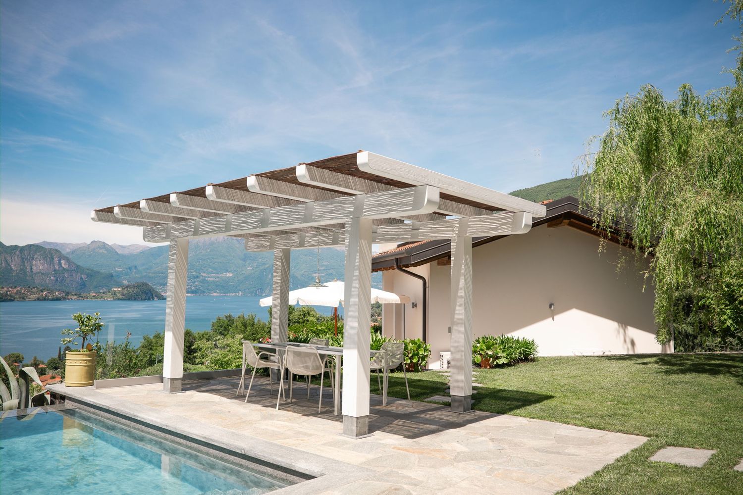 White pergola with outdoor dining set beside a pool, overlooking a serene lake and mountains under a blue sky.