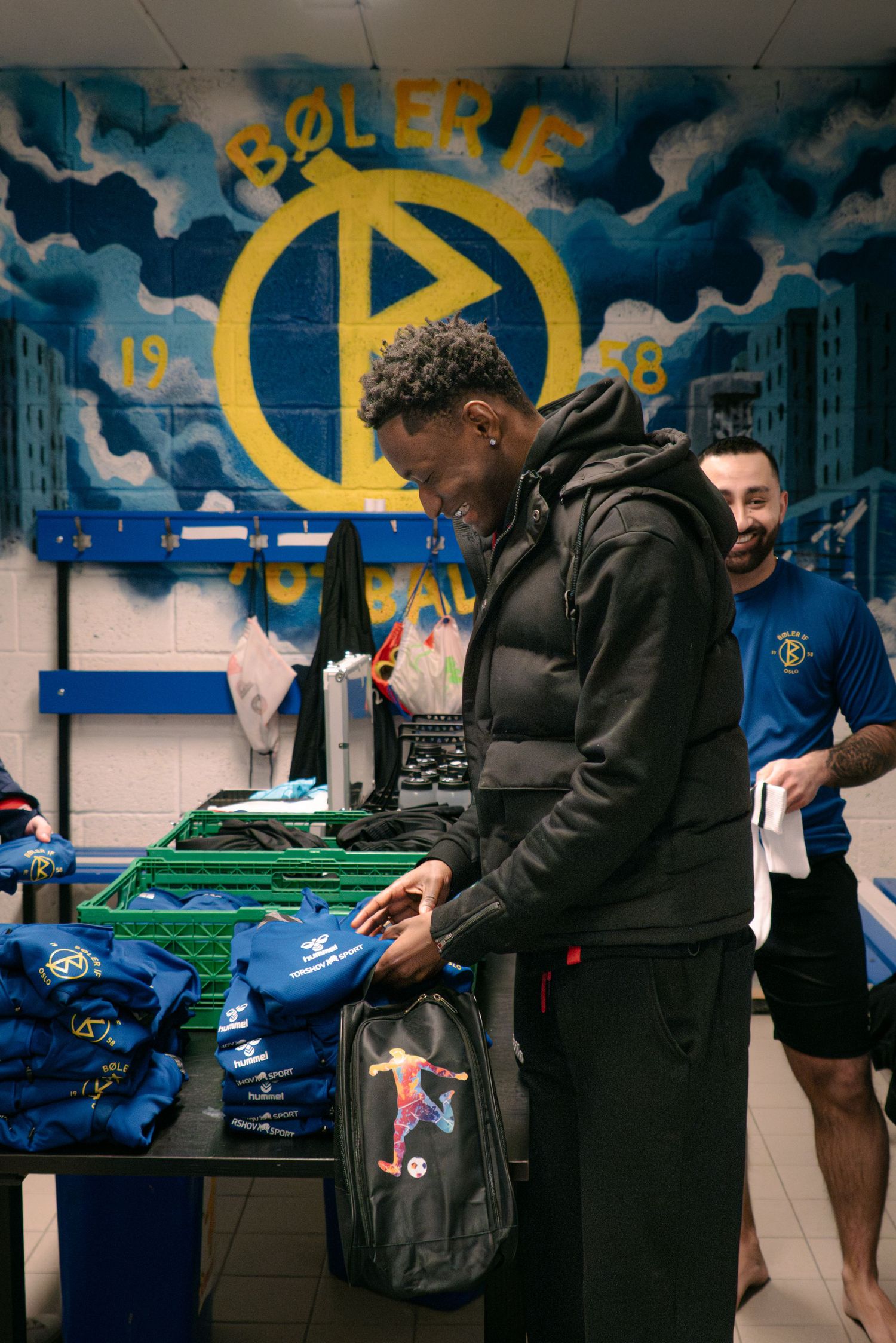 Man in black hoodie unpacking sports gear in a locker room with Bøler IF club mural on the wall.