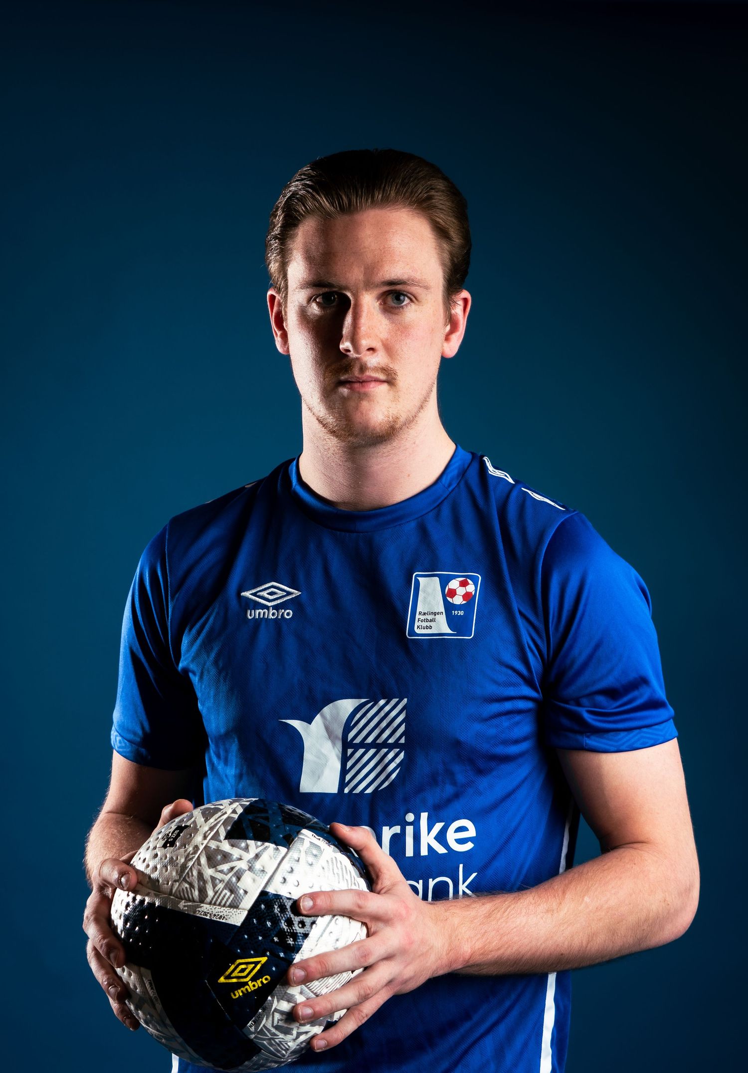 Athlete in blue uniform holds silver sports equipment against dark studio backdrop.