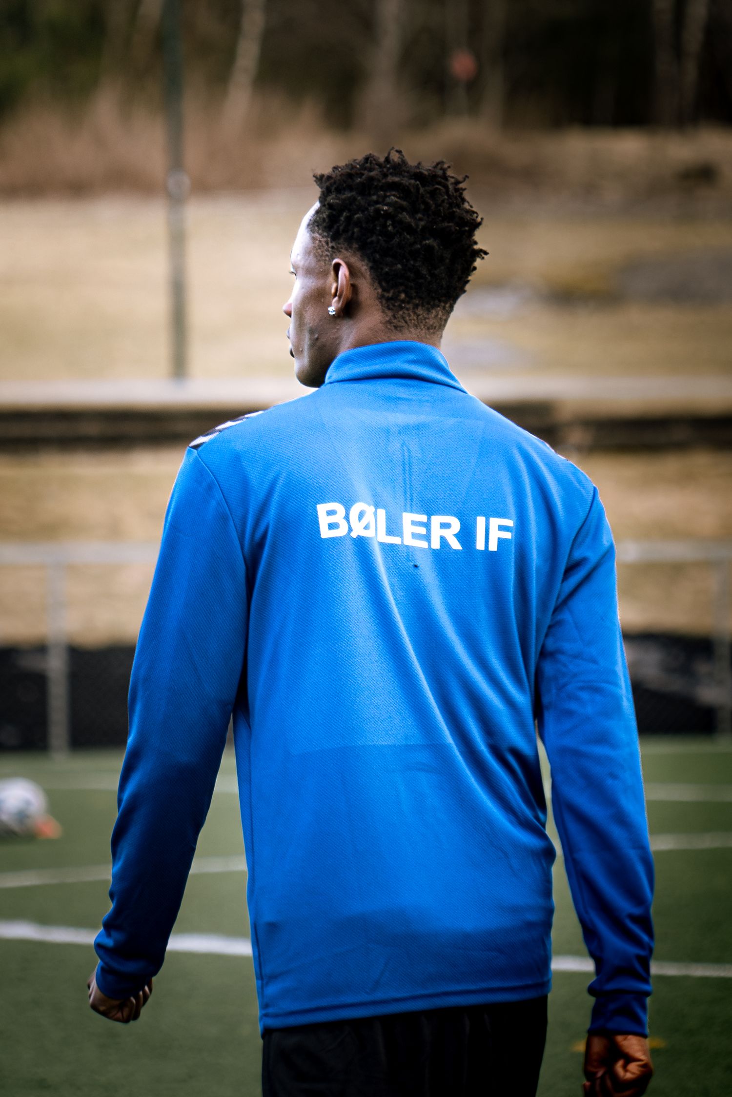 Man wearing blue Boler IF sports jacket standing on football pitch, viewed from behind.