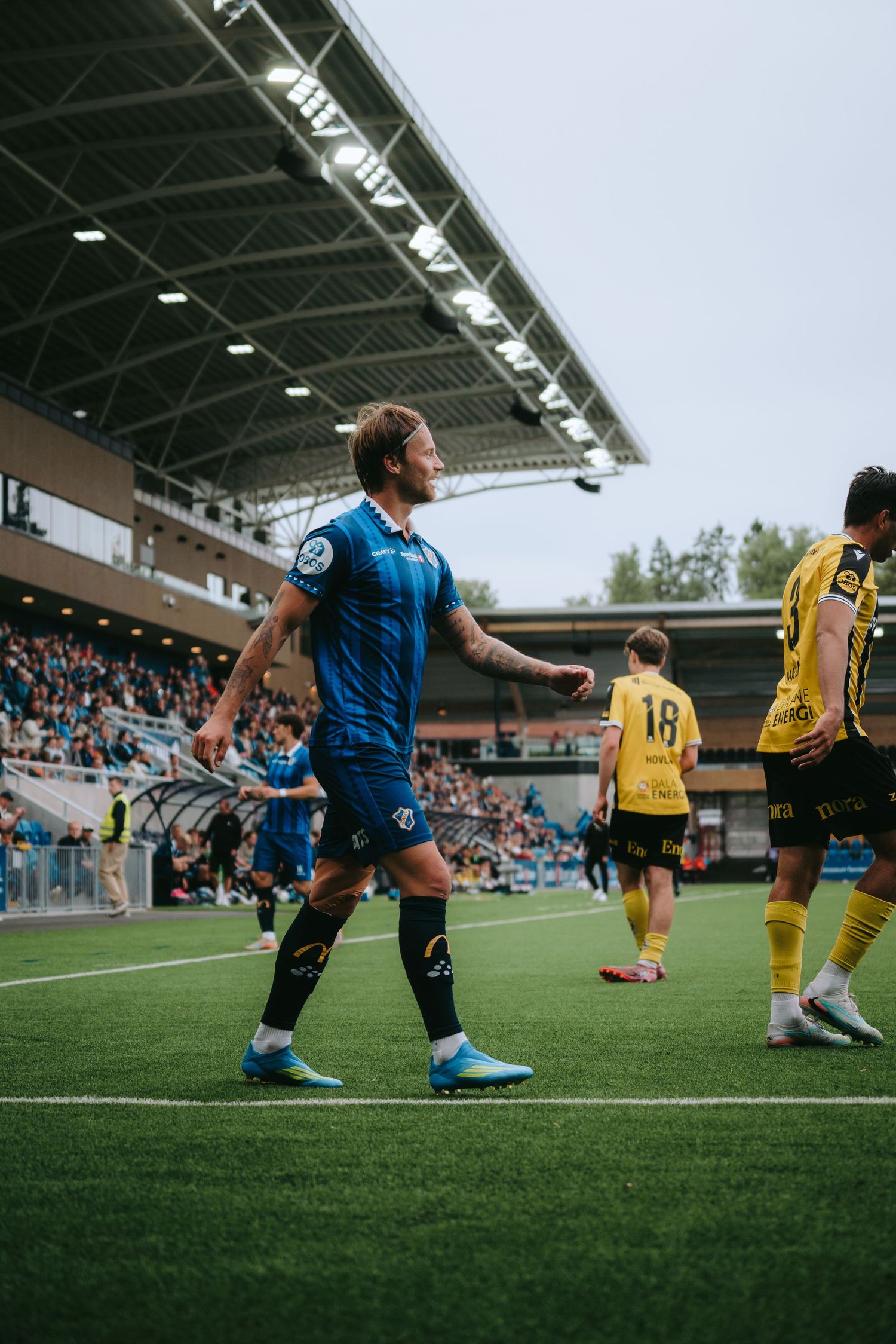 Soccer players in blue and yellow jerseys compete on a green field in a modern stadium during a daytime match.