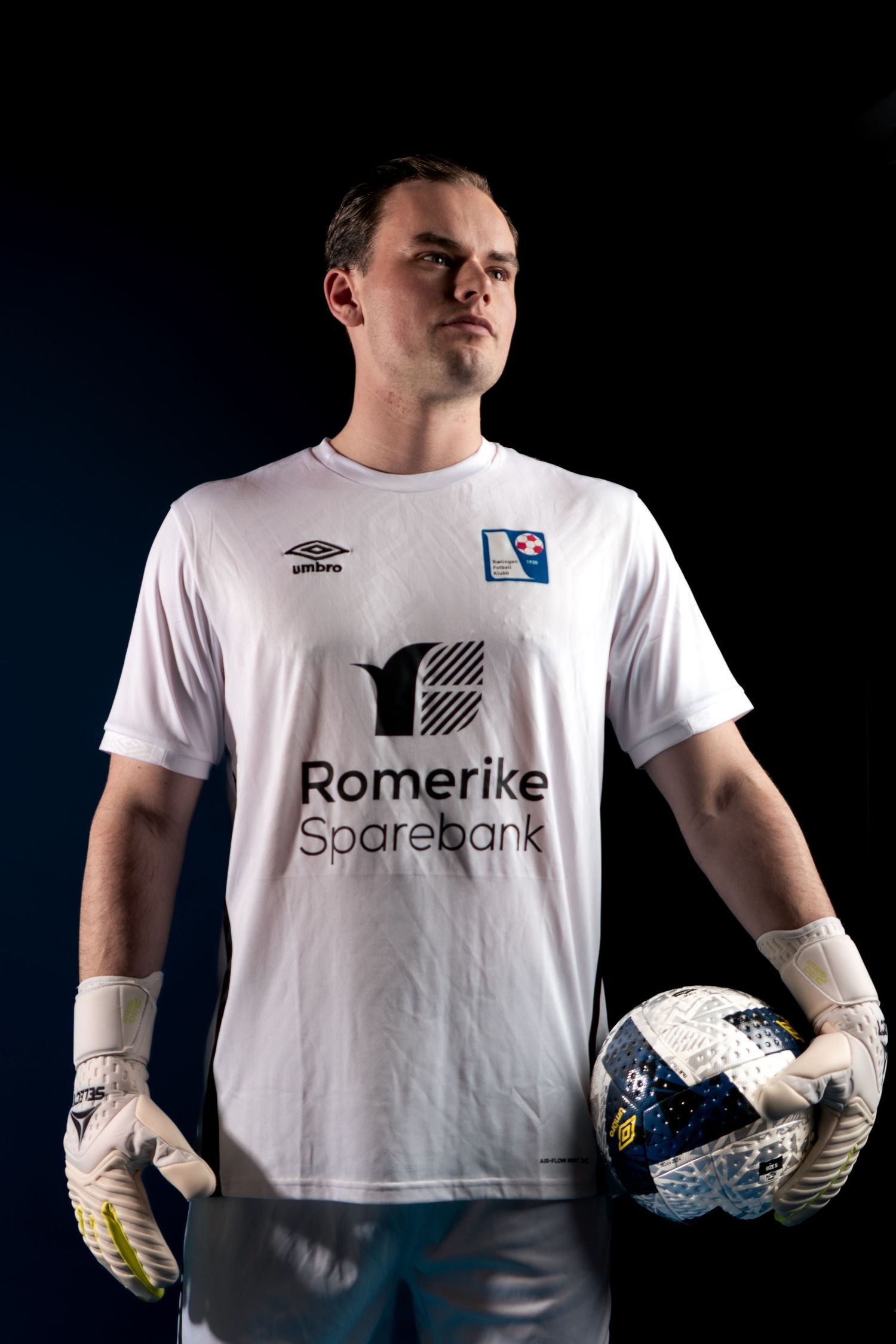Soccer goalkeeper in white Romerike jersey holding ball against dark background.