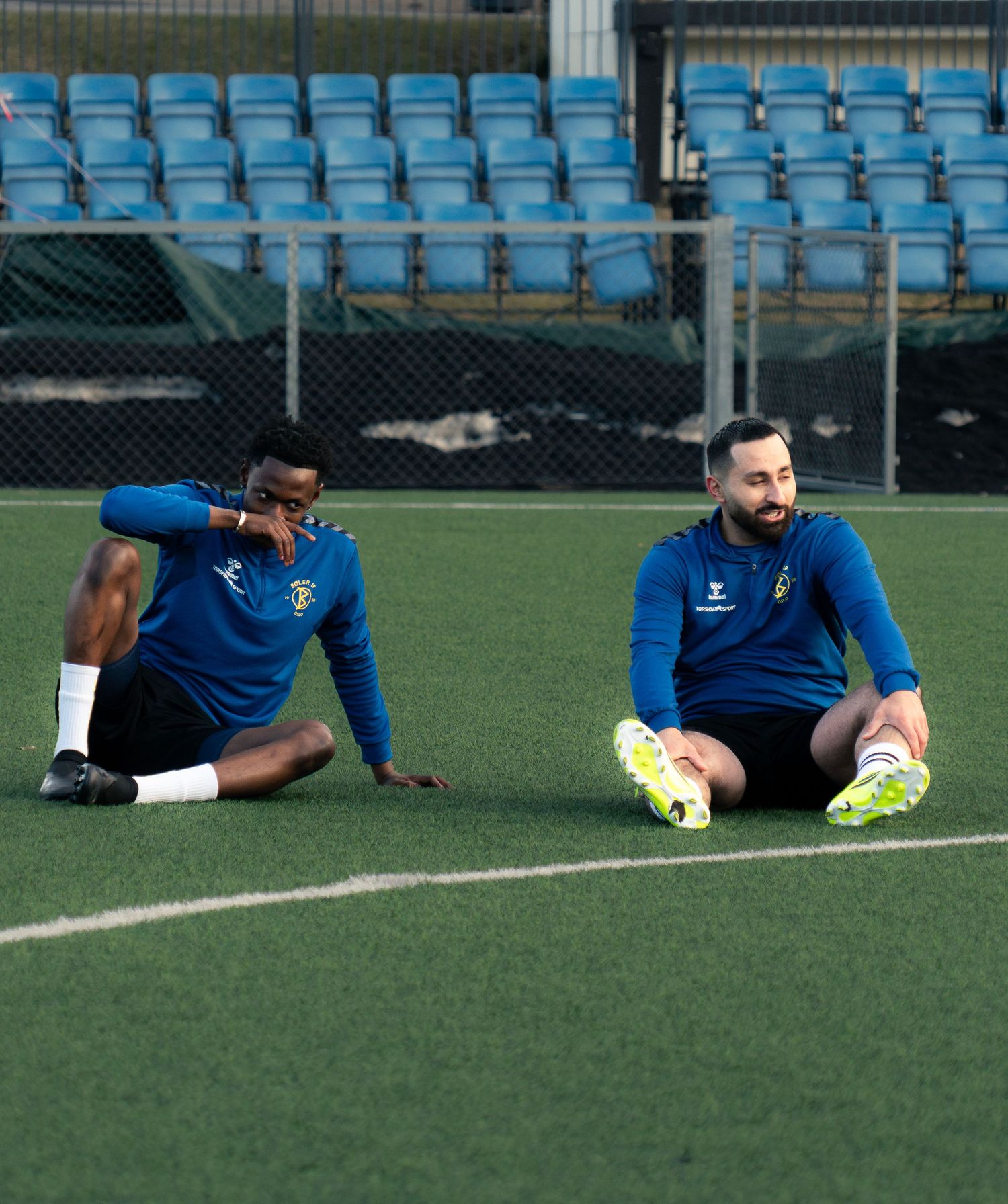 Two soccer players in blue kits stretch on a green turf field with blue stadium seats in the background.