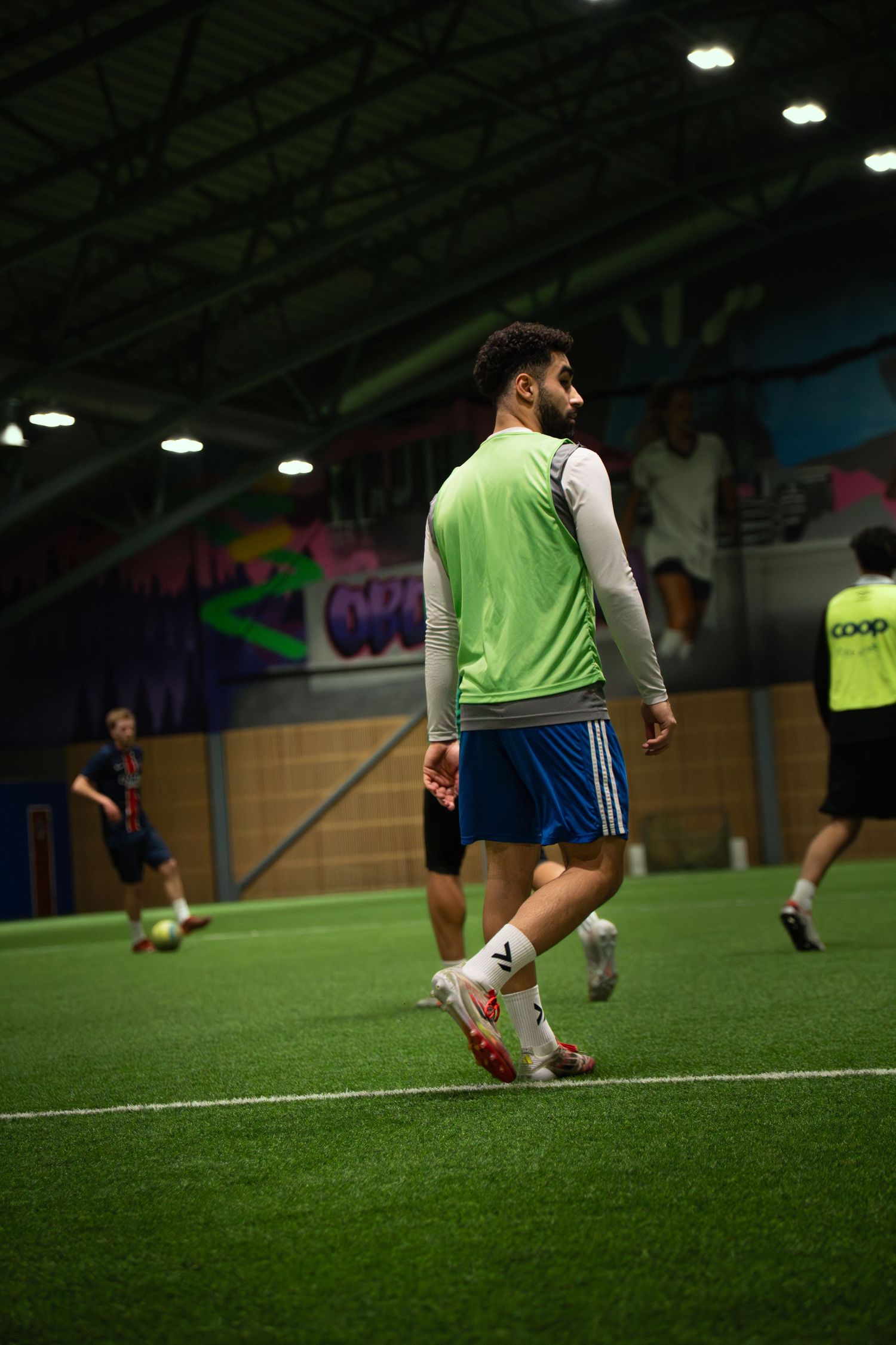 Players compete in an indoor futsal match on artificial turf, wearing colorful bibs under bright arena lighting.