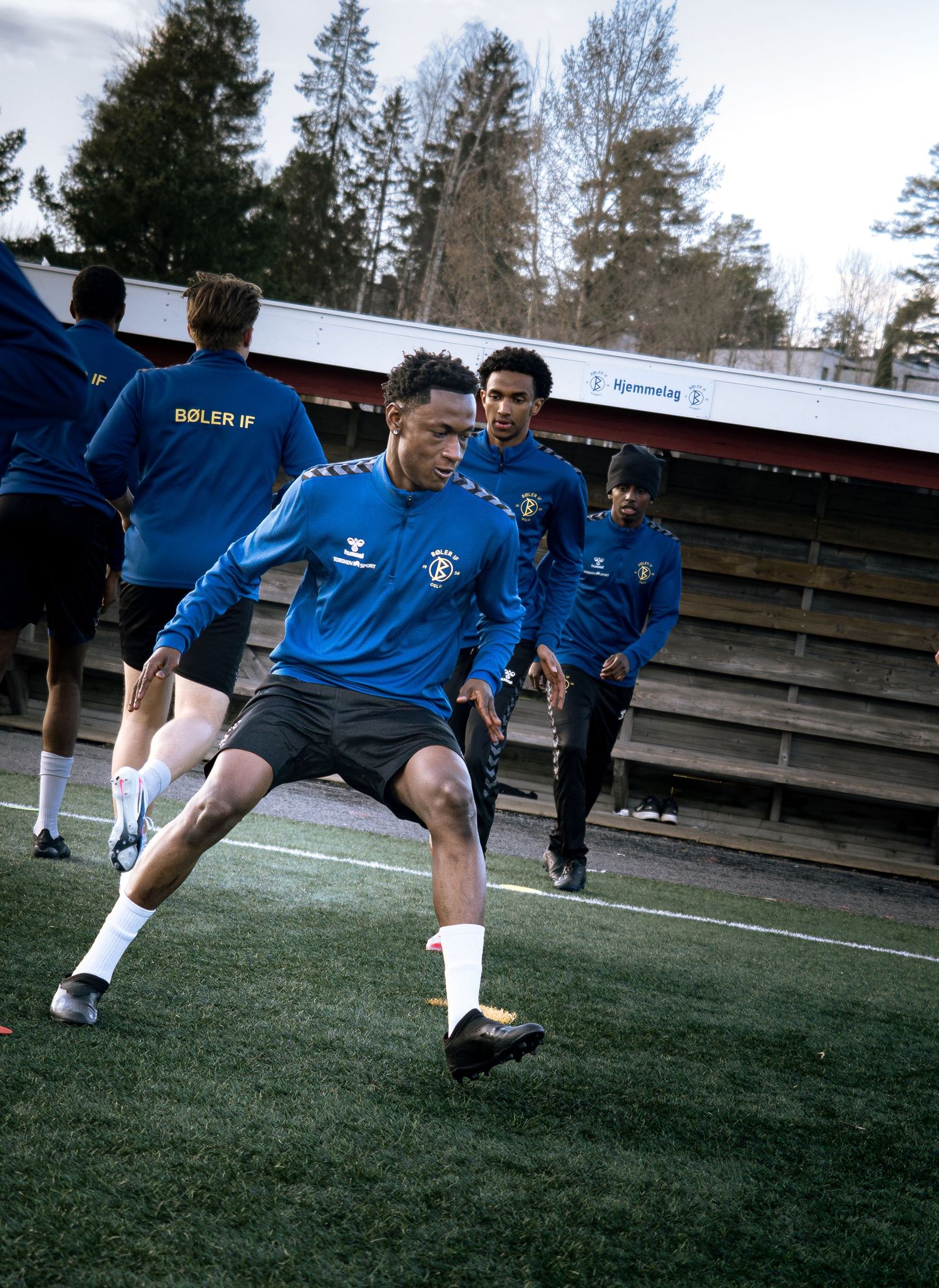 Soccer players in blue Hummel jerseys practice drills on an outdoor turf field surrounded by trees.